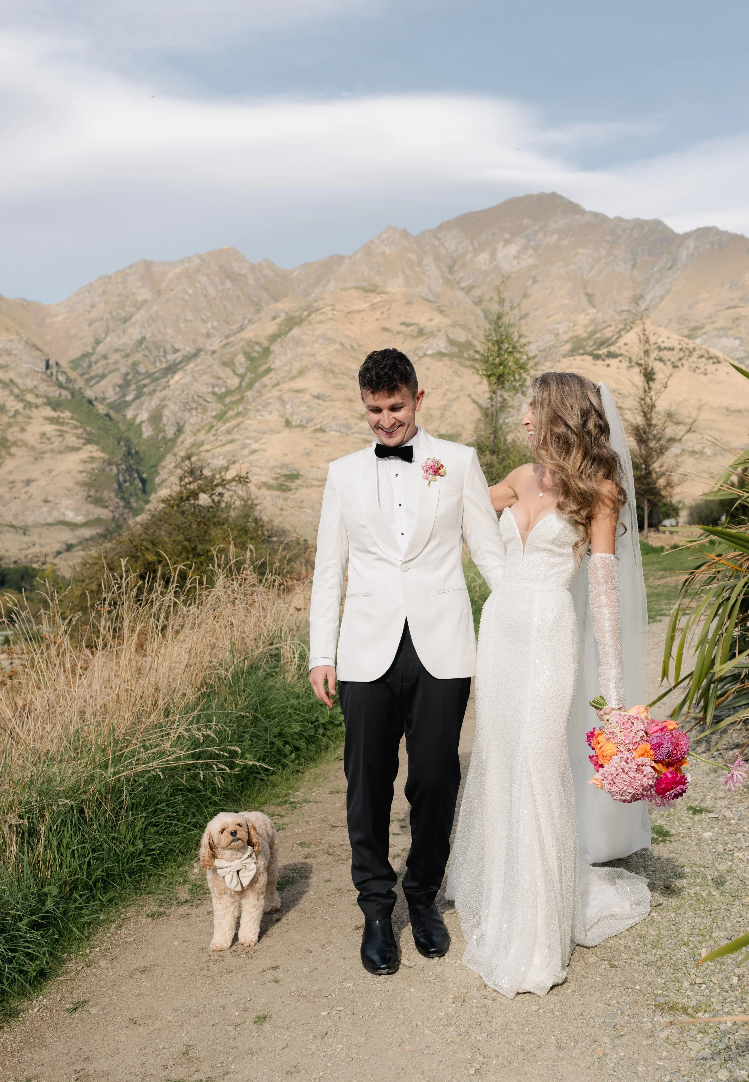 A bride and groom in wedding attire walking outdoors on a dirt path, smiling and holding hands, with a small dog nearby, mountains in the background, dark clouds in the sky, and greenery around.