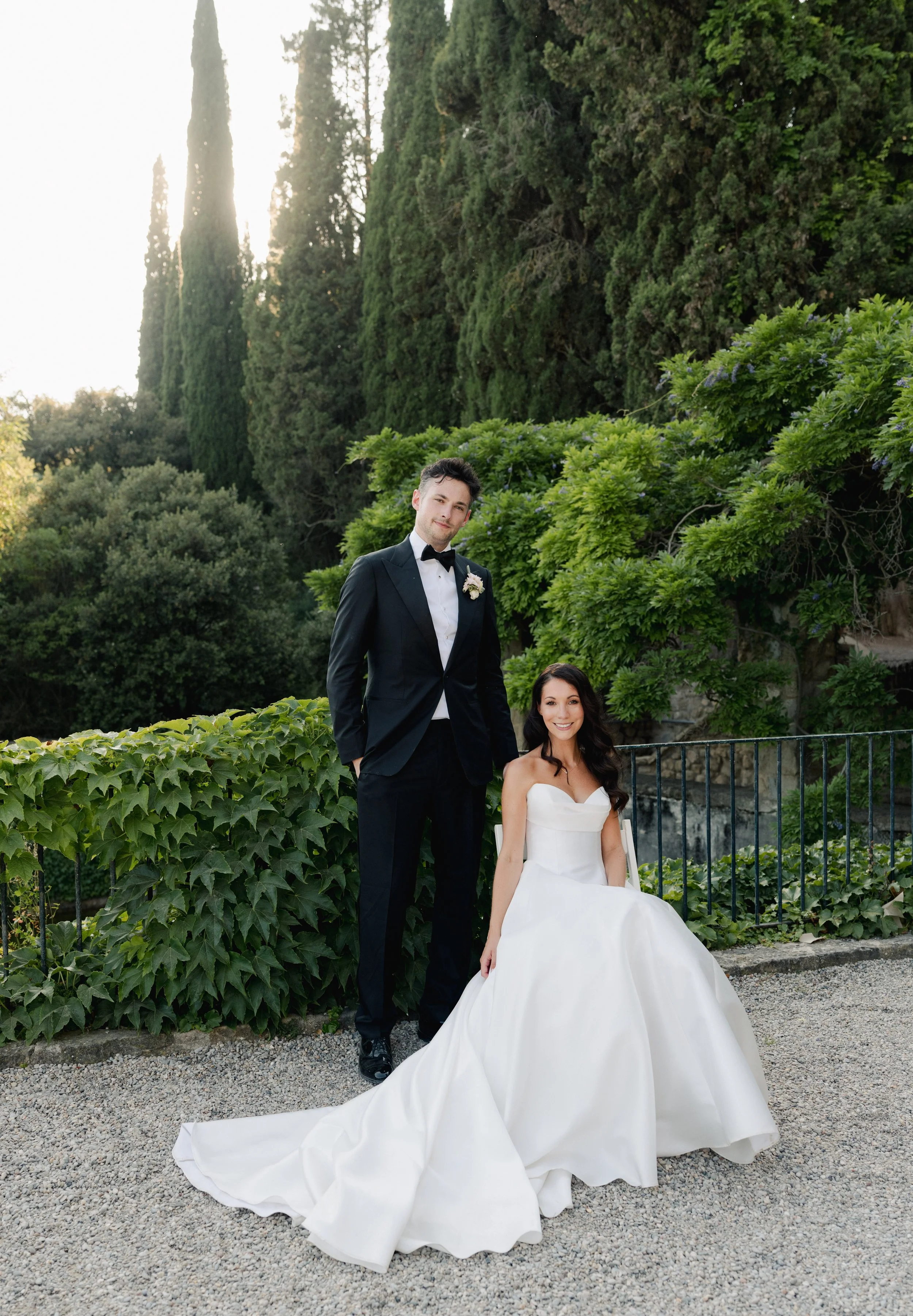 A bride in a white wedding gown sitting on the ground and a groom in a black tuxedo standing beside her in an outdoor garden setting.