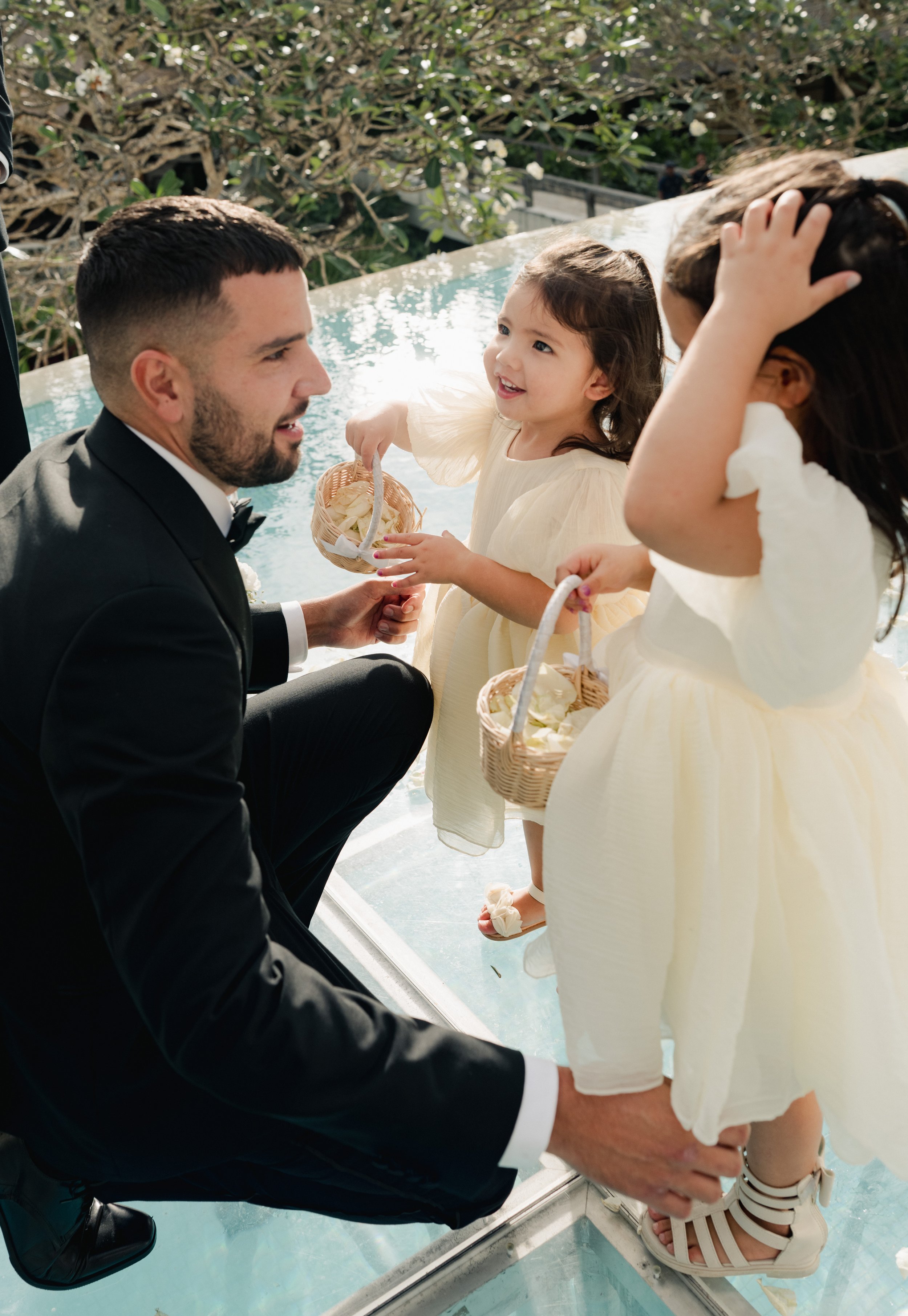 A man and two young girls dressed in light yellow dresses holding small baskets of flower petals, with the man crouching and smiling at the girls, on a glass platform by a pool, outdoors with greenery in the background.