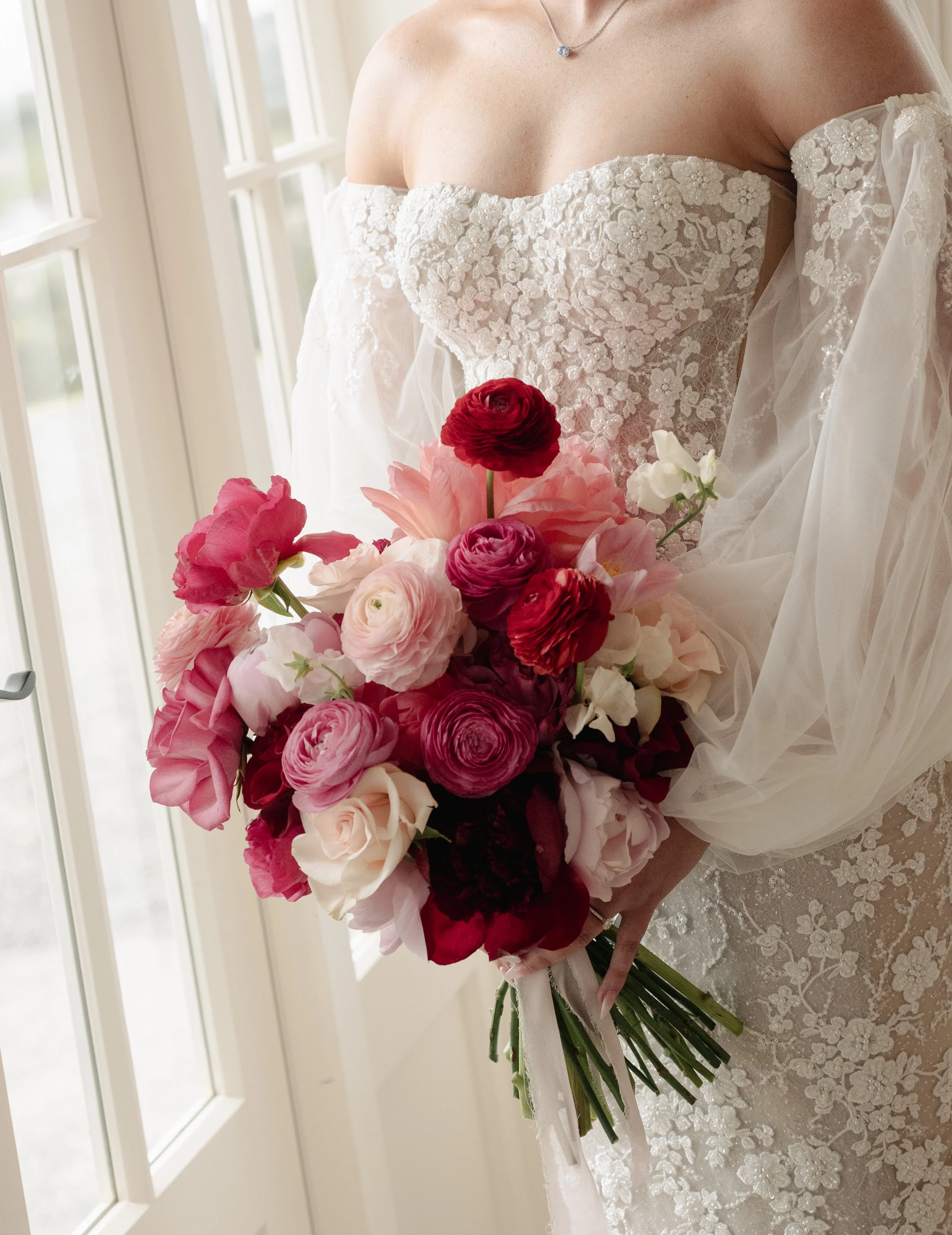 A bride in a lace off-shoulder wedding dress holding a bouquet of pink, red, white, and dark burgundy flowers near a window.
