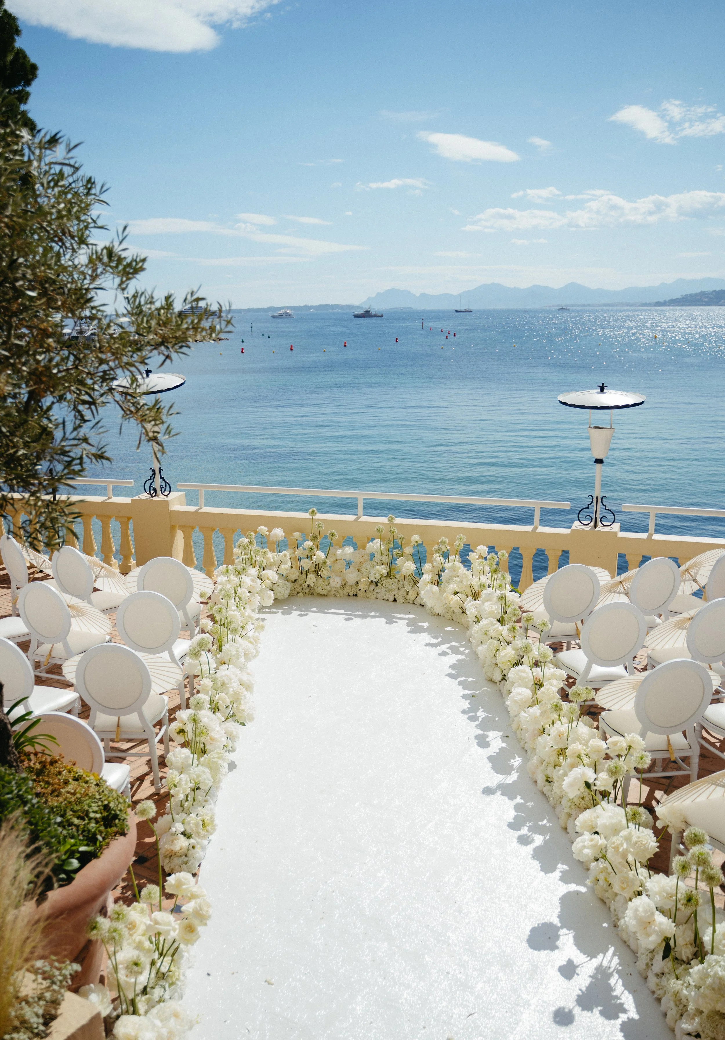 Outdoor wedding altar with white flower arrangements along an aisle, overlooking a calm ocean with boats and mountains in the background on a bright, sunny day.