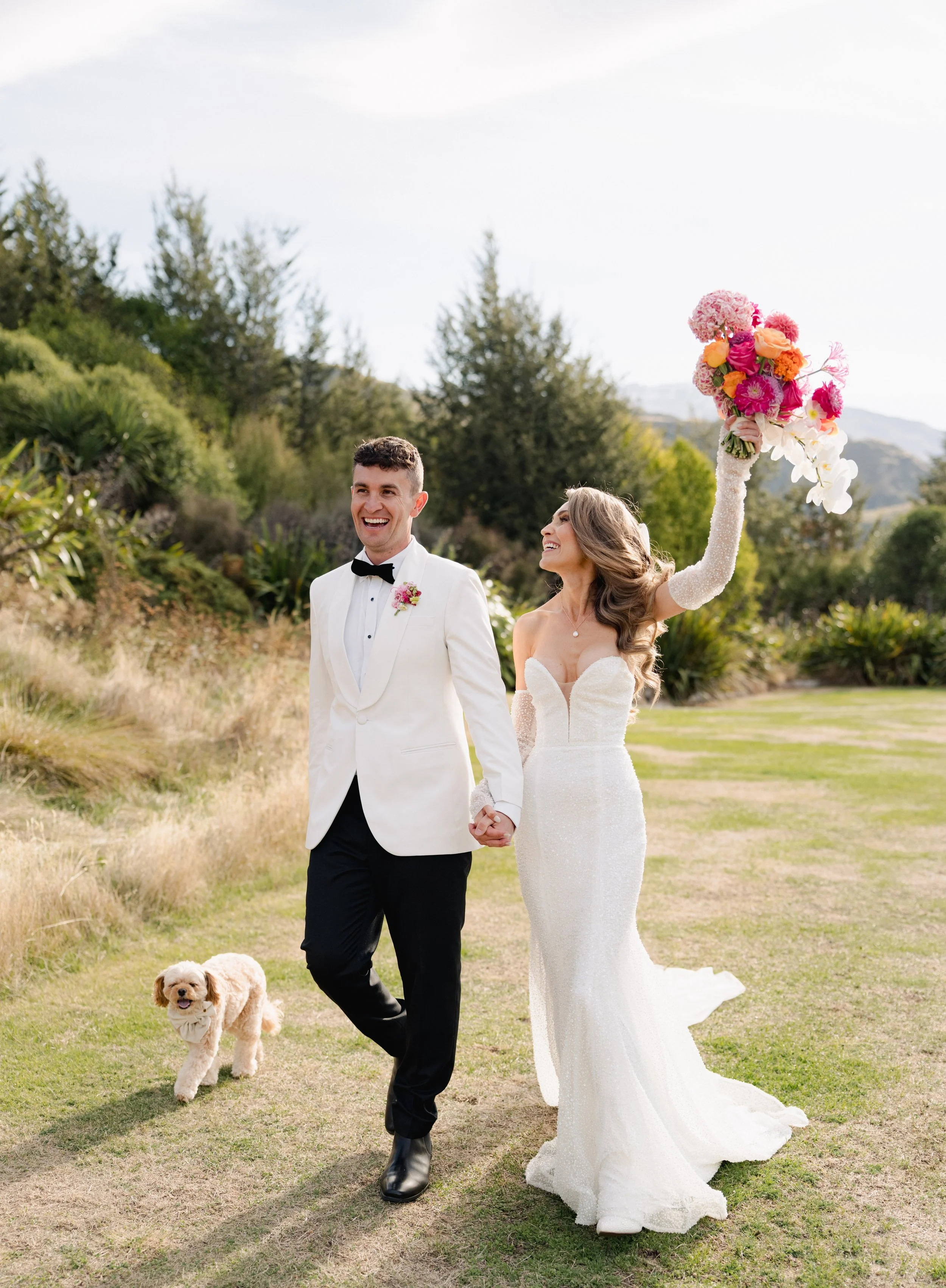 A bride and groom walking hand in hand outdoors on their wedding day. The bride is holding a large bouquet of pink, orange, and white flowers and has a joyful expression. The groom is dressed in a white tuxedo jacket, black pants, and a black bow tie