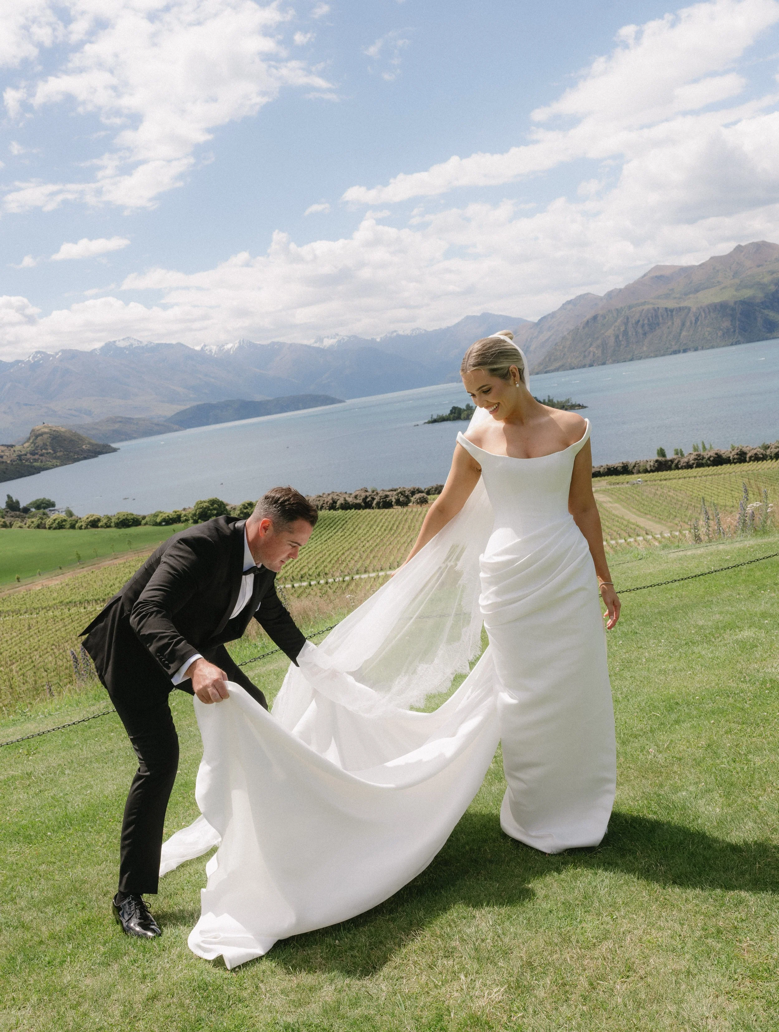 A bride and groom, outdoors by a lake, with mountains in the background. The groom is lifting the bride's train as she smiles.