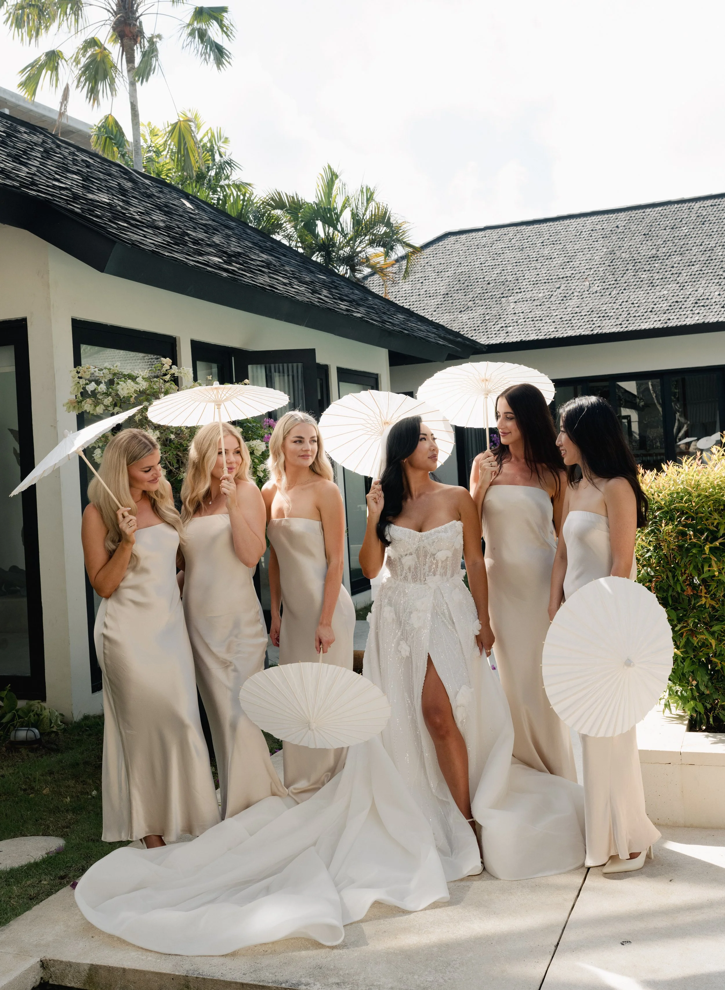 Bride and five bridesmaids holding white umbrellas outside a modern house with tropical plants.