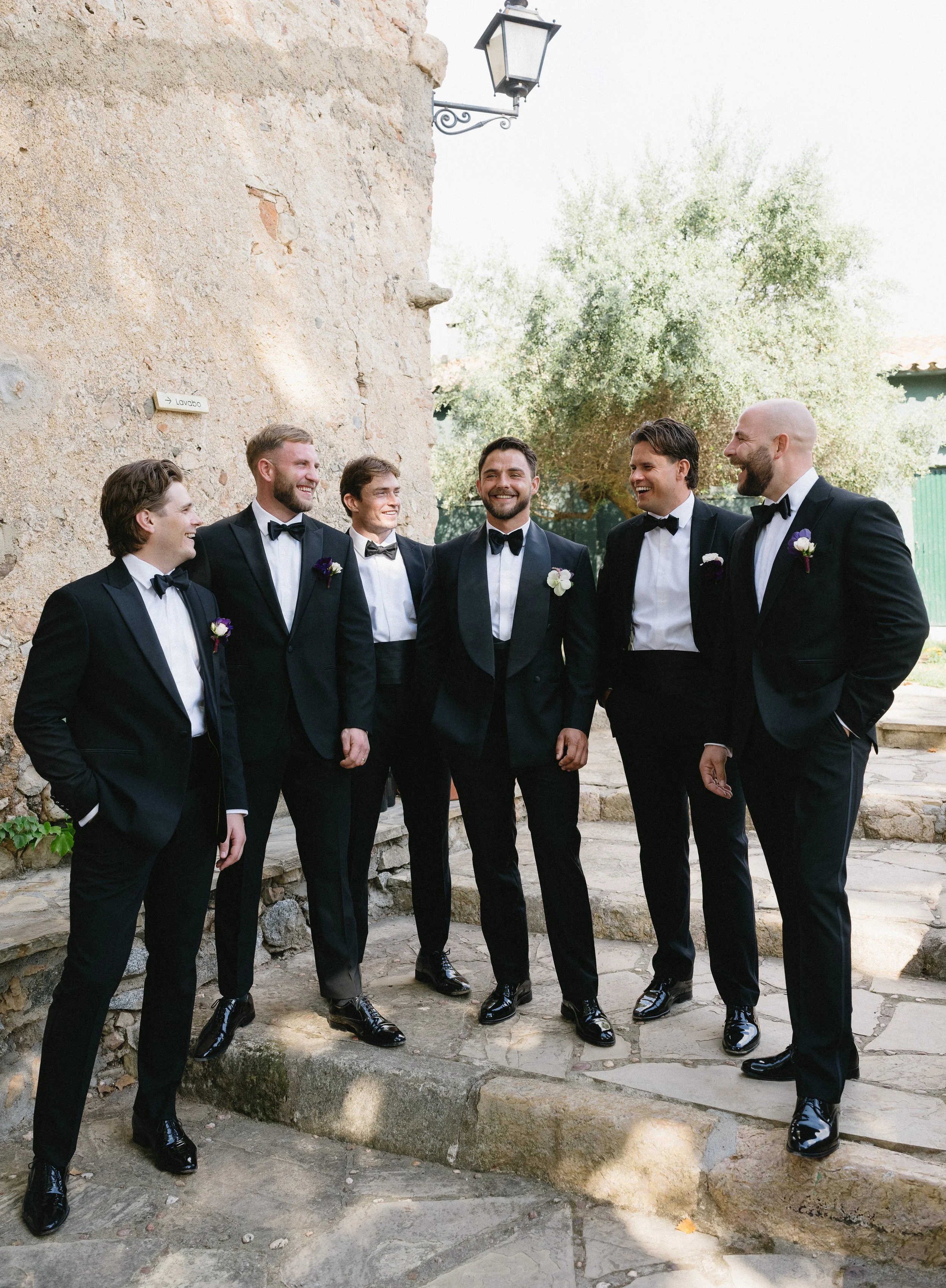 Seven men dressed in black tuxedos with bow ties and boutonnières, standing outdoors on a stone pathway near a rustic stone wall and greenery, smiling and talking together.