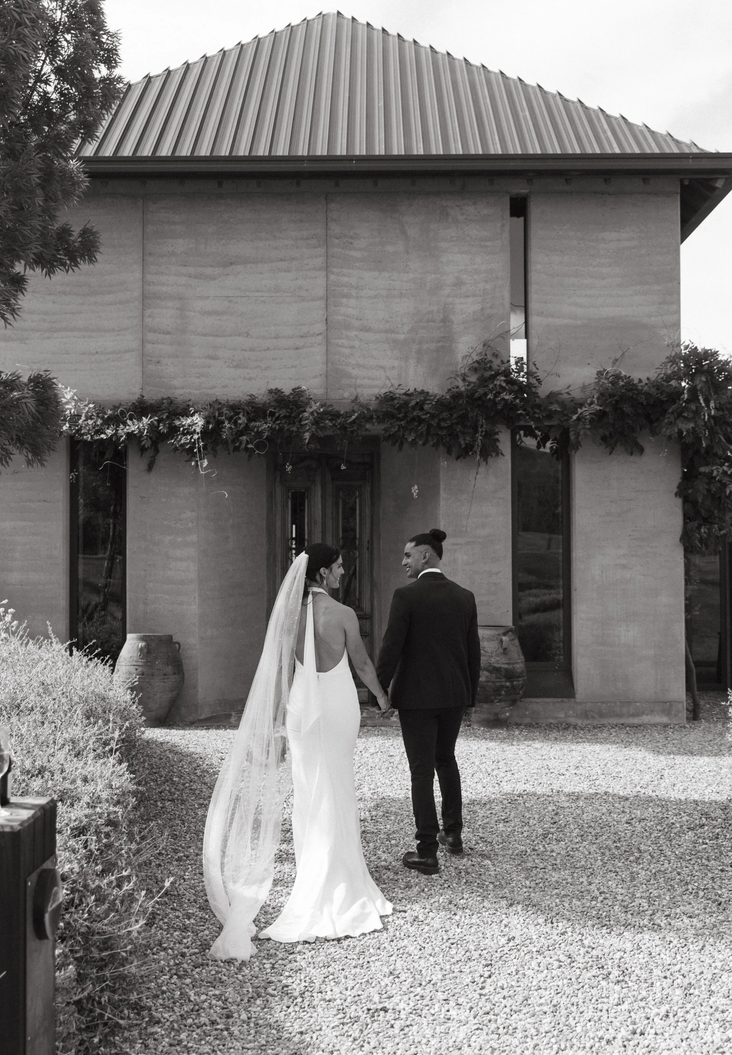 Black and white photo of a bride and groom holding hands outside a modern building, dressed in wedding attire, with the bride in a long white gown and veil, and the groom in a dark suit, under hanging greenery.