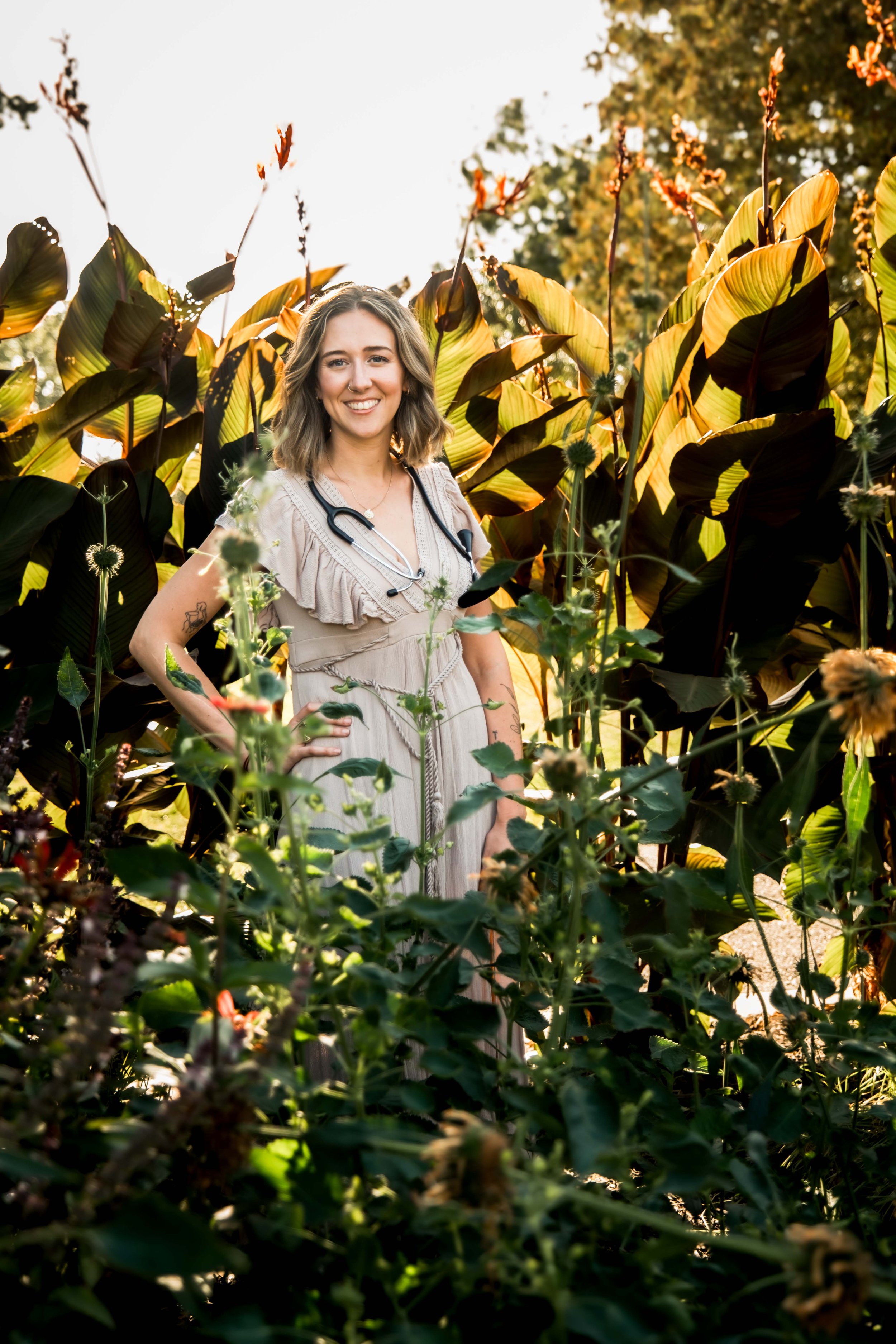 Student midwife Lauren Maggi standing in a garden with lush greenery behind her and flowers in front of her. She is wearing a cream colored dress, has a hand on her hip and a fetoscope around her neck.