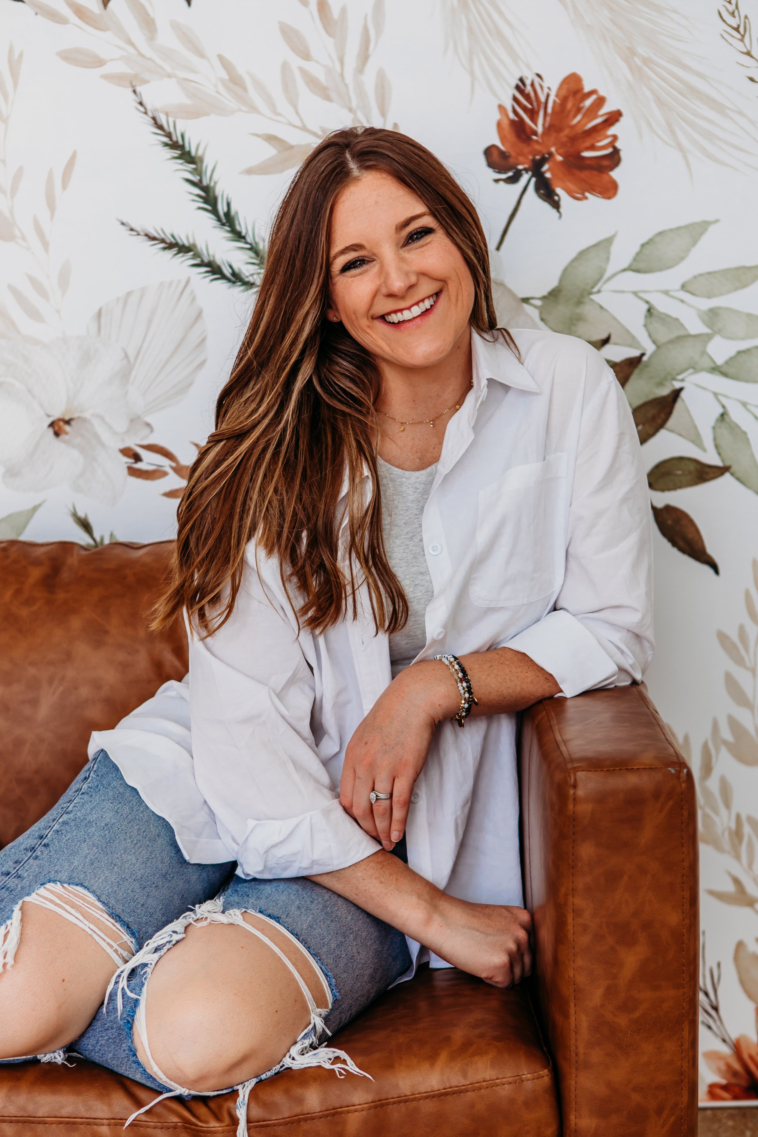 Tess is sitting indoors on a brown couch with a floral background. She's wearing white shirt and jeans with holes in the knees