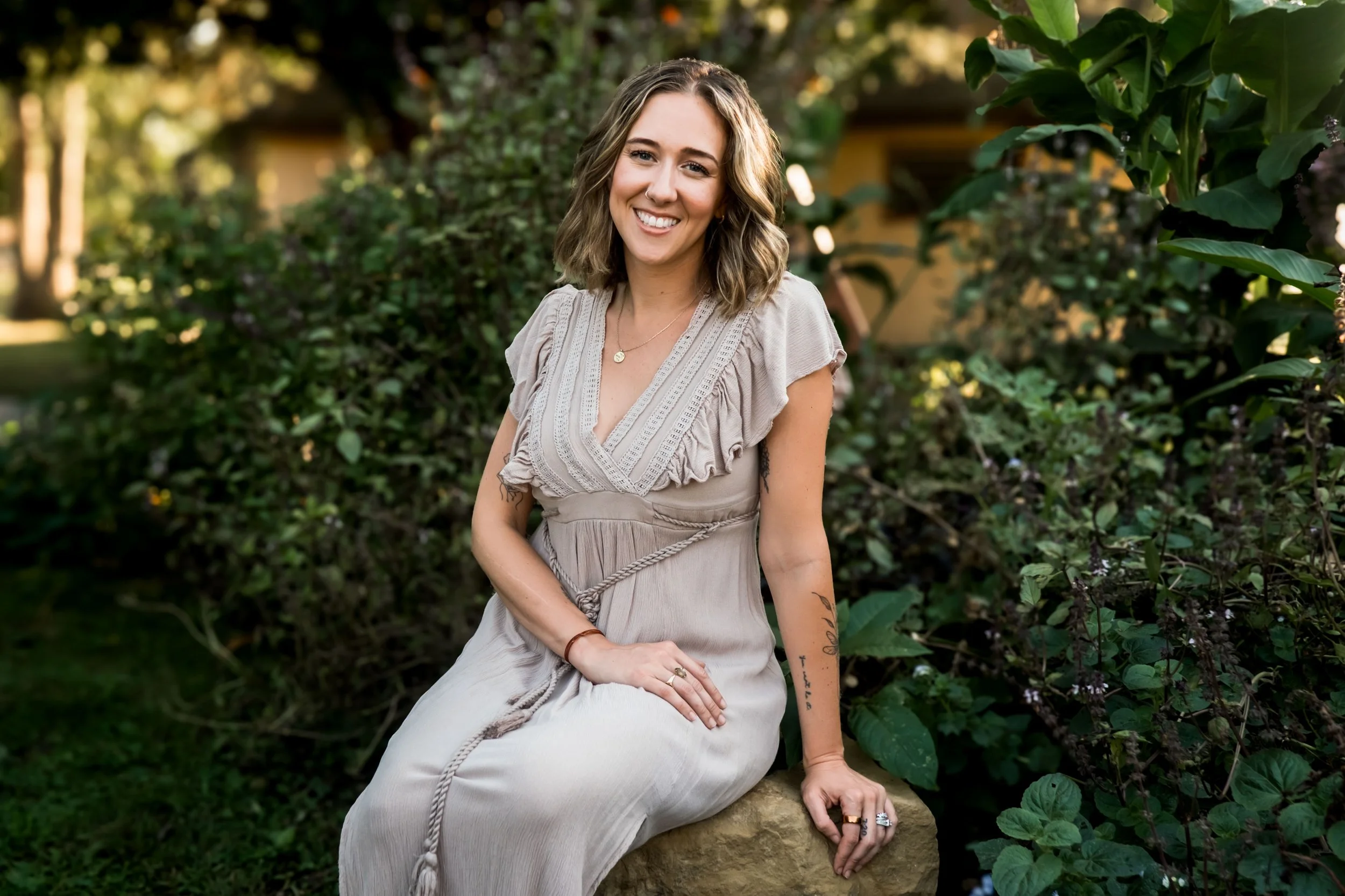 Student Lauren Maggi in a cream colored dress sitting outside on a rock, lush greenery in the background. She has small tattoos on both arms.