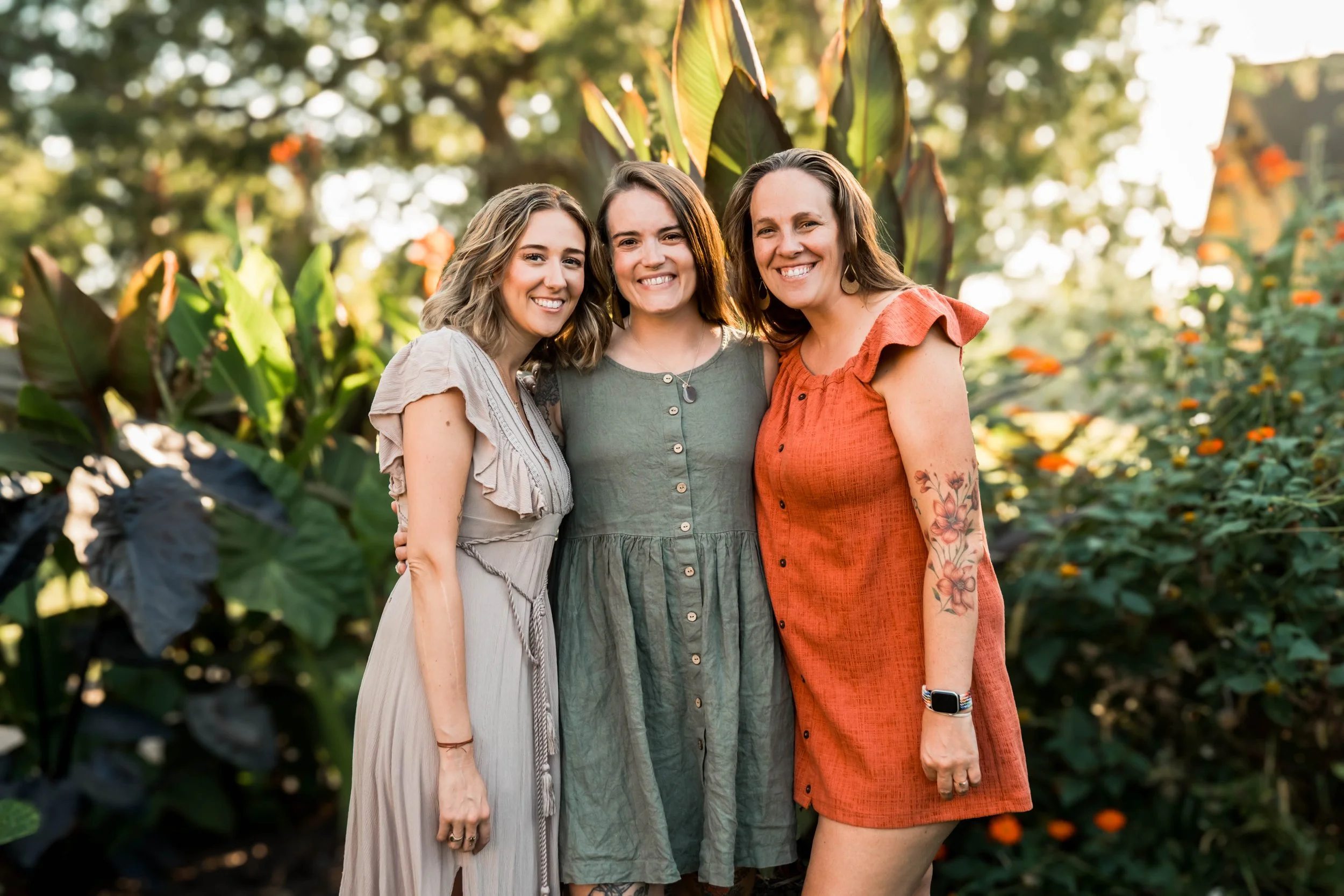 Lauren Maggi (cream dress), Sarah Moore (green dress), and Steph Thoms (orange dress) stand in a garden, arms around each other. Lush greenery in the background.