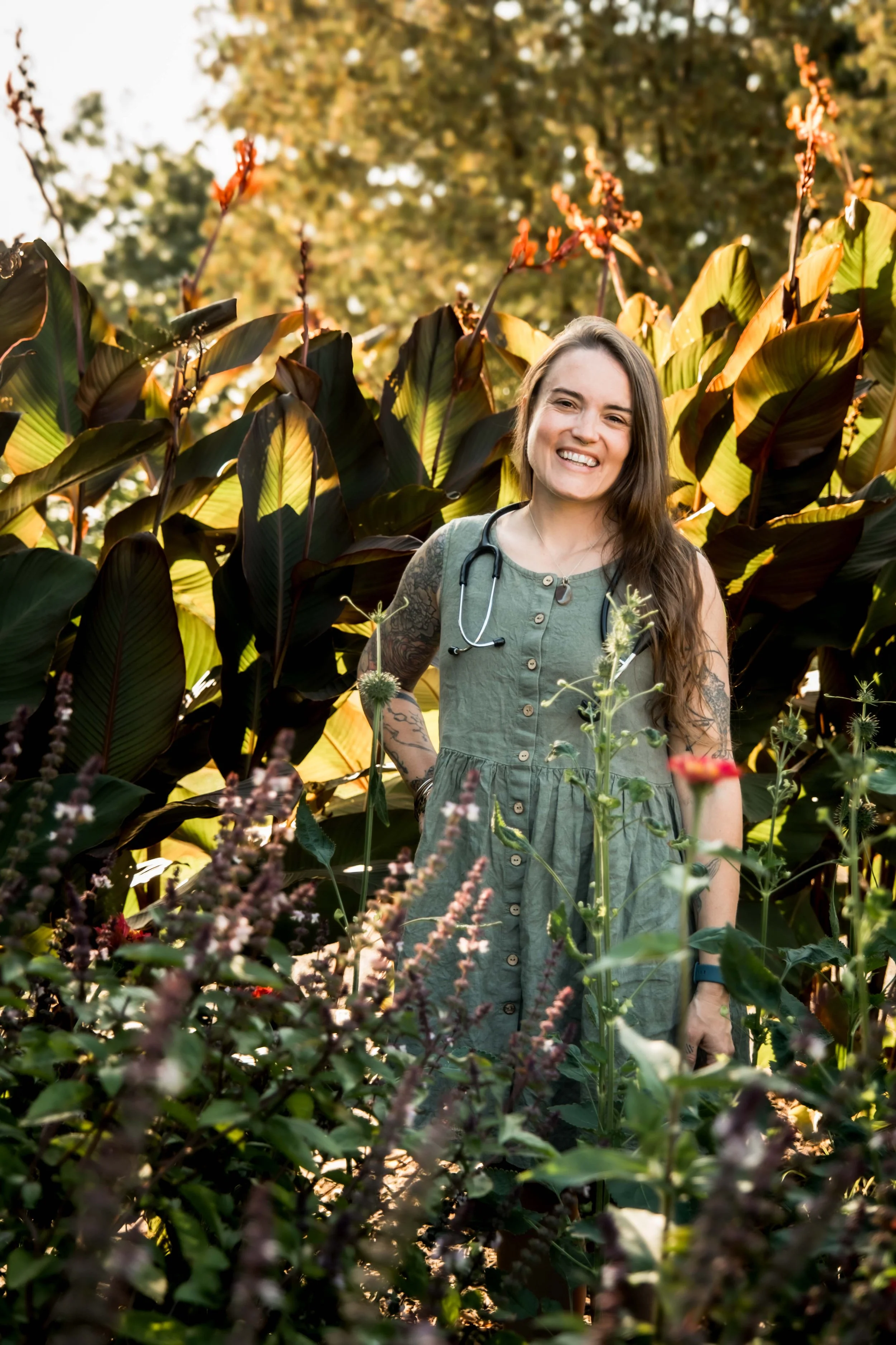 Midwife Sarah Moore standing in a garden with lush plants behind her and flowers in front of her. She has tattoos on her arms and a fetoscope around her neck.