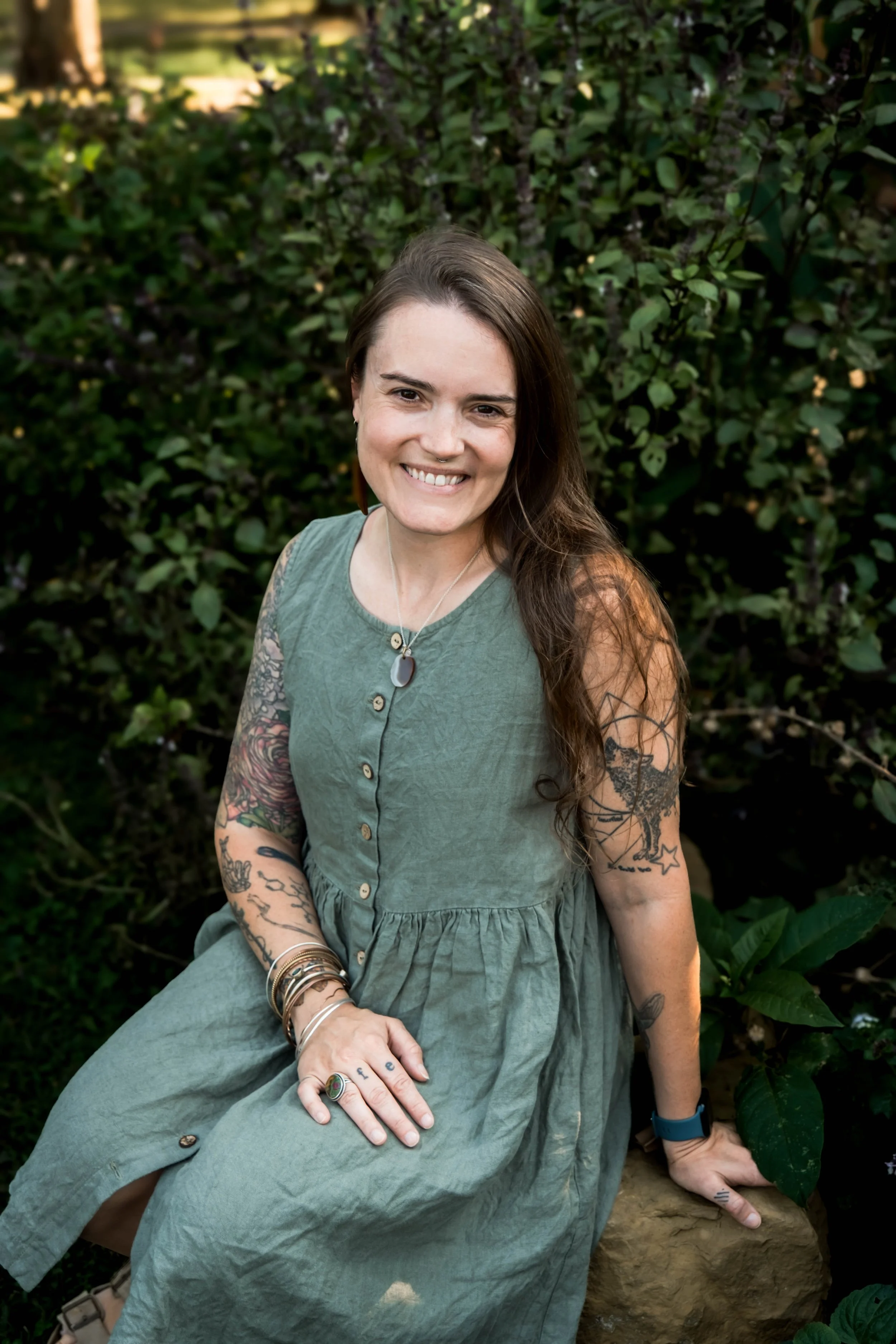 Midwife Sarah Moore wearing a sage green dress with buttons. She's sitting outdoors with lush greenery in the background. She has tattoos on her arms and long hair.