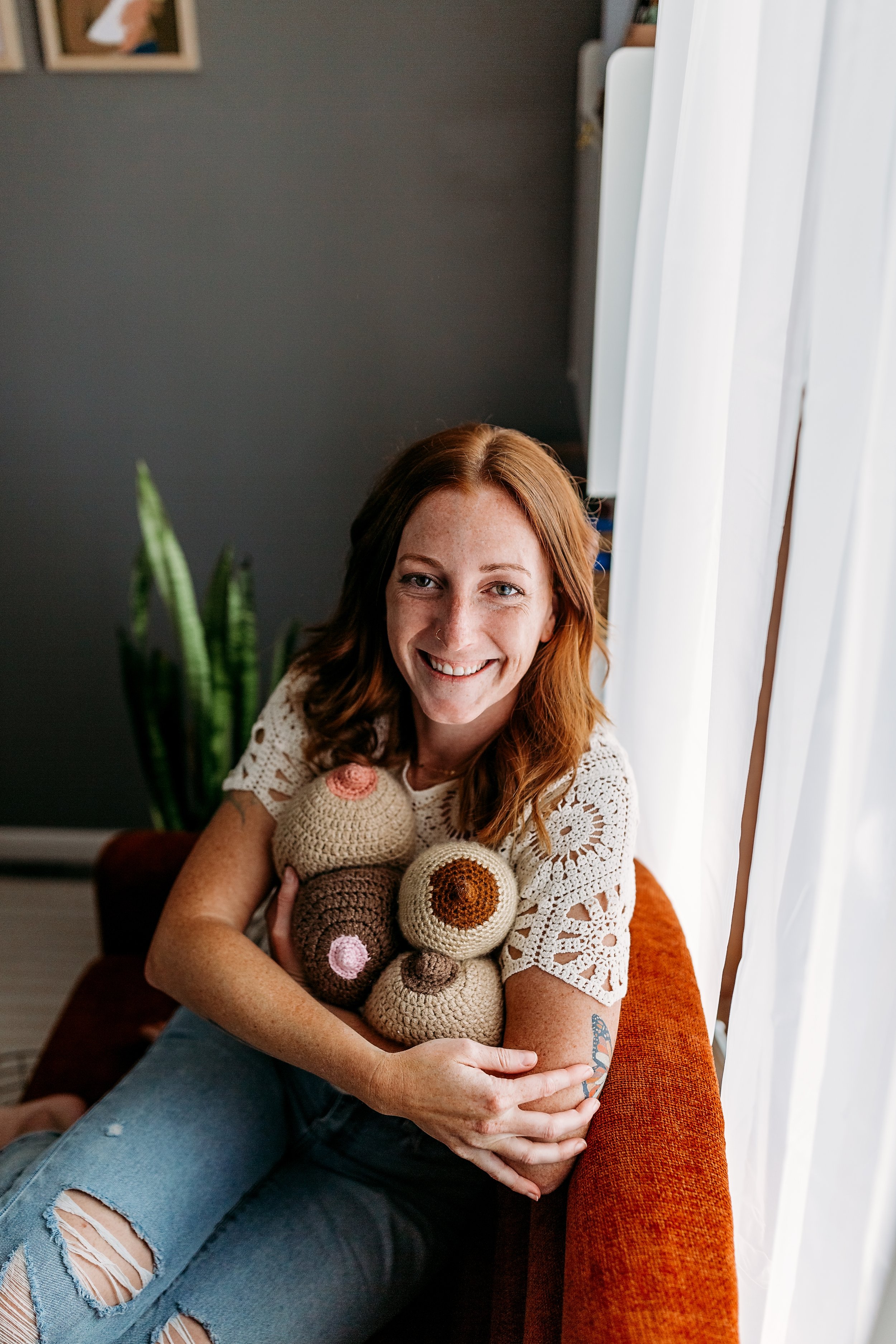 Kenzie is sitting on an orange couch with a gray background. She's wearing a white crocheted top and holding an arm full of crocheted breasts!