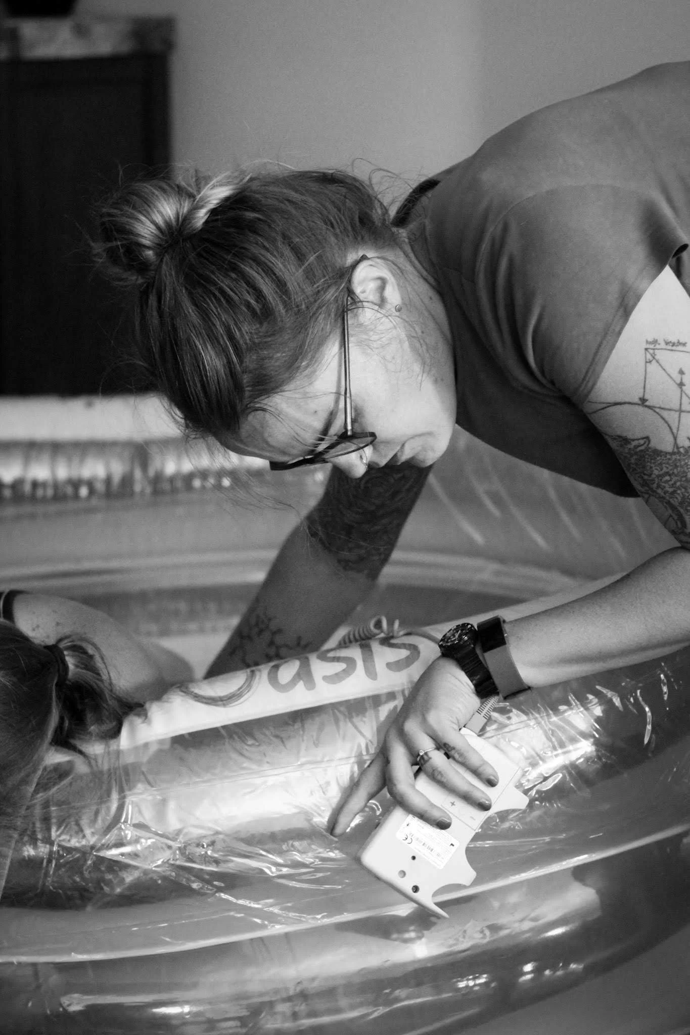 Midwife Sarah Moore, black and white photo, leaning over with a doppler to auscultate fetal heart tones. Her client is visible to the left, in a birth pool that says "Oasis." Sarah has tattoos on her arms, a watch and a bracelet on, and glasses.