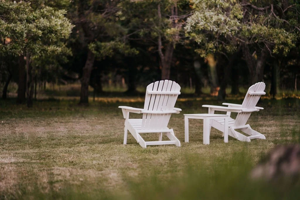 Two white wooden deck chairs sit on the lawn, surrounded by bush