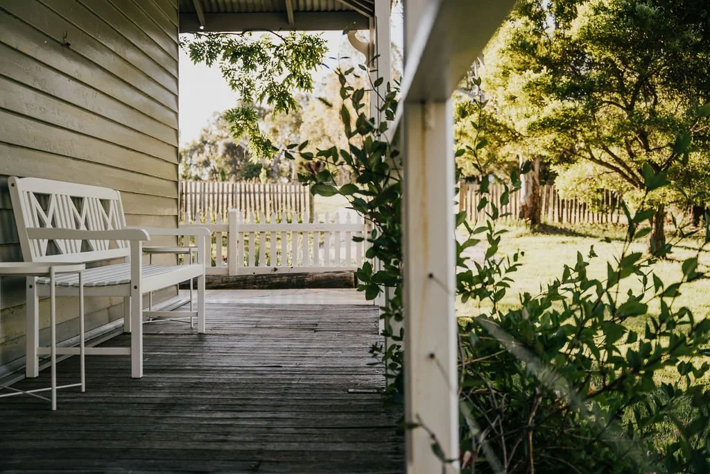 A white bench seat on a cottage verandah with late afternoon light in the background