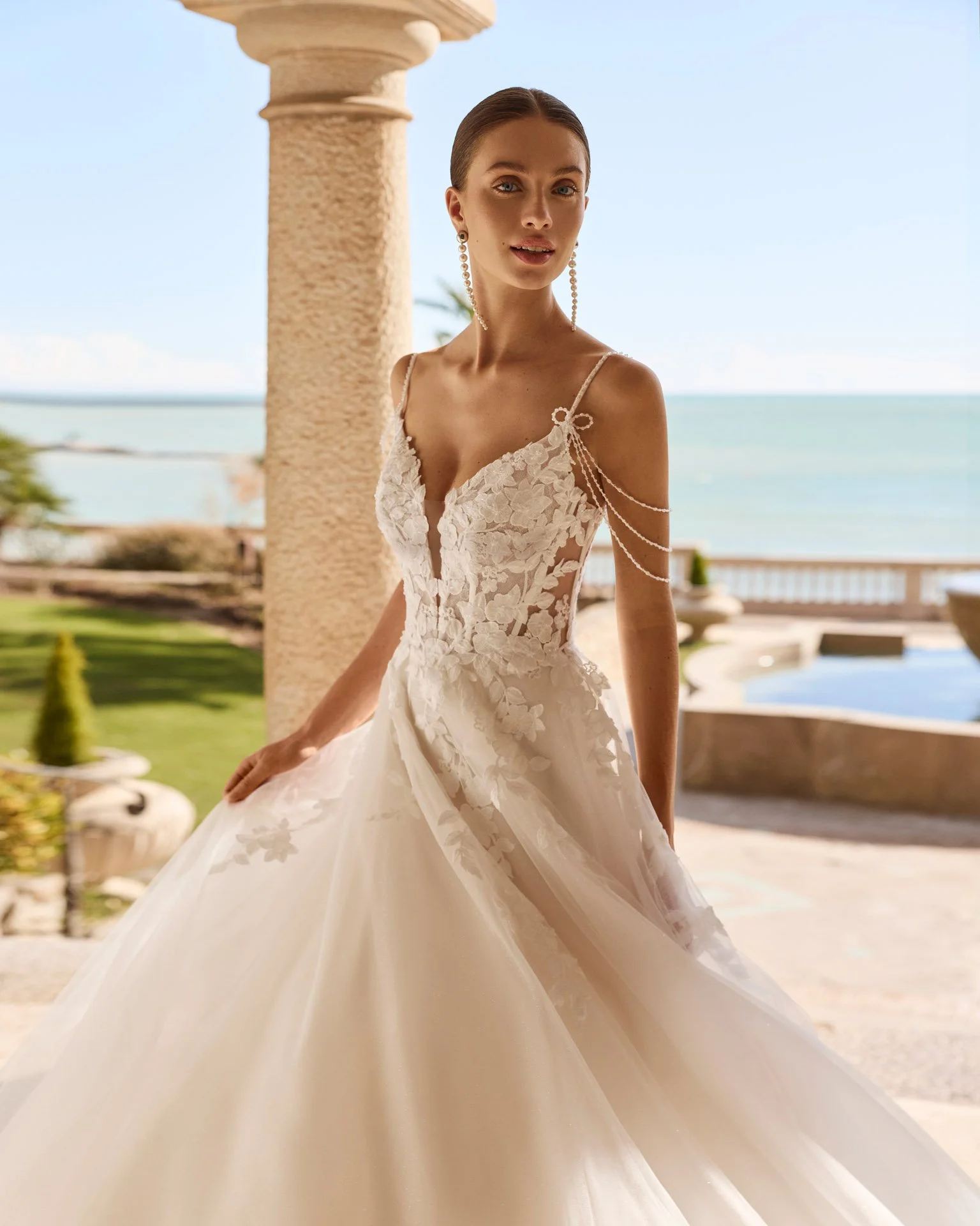Vestido de noiva Flash com decote em V profundo, rendas delicadas, transparências laterais, alças finas em pedraria, laços e fios de pedraria caídos sobre os ombros.