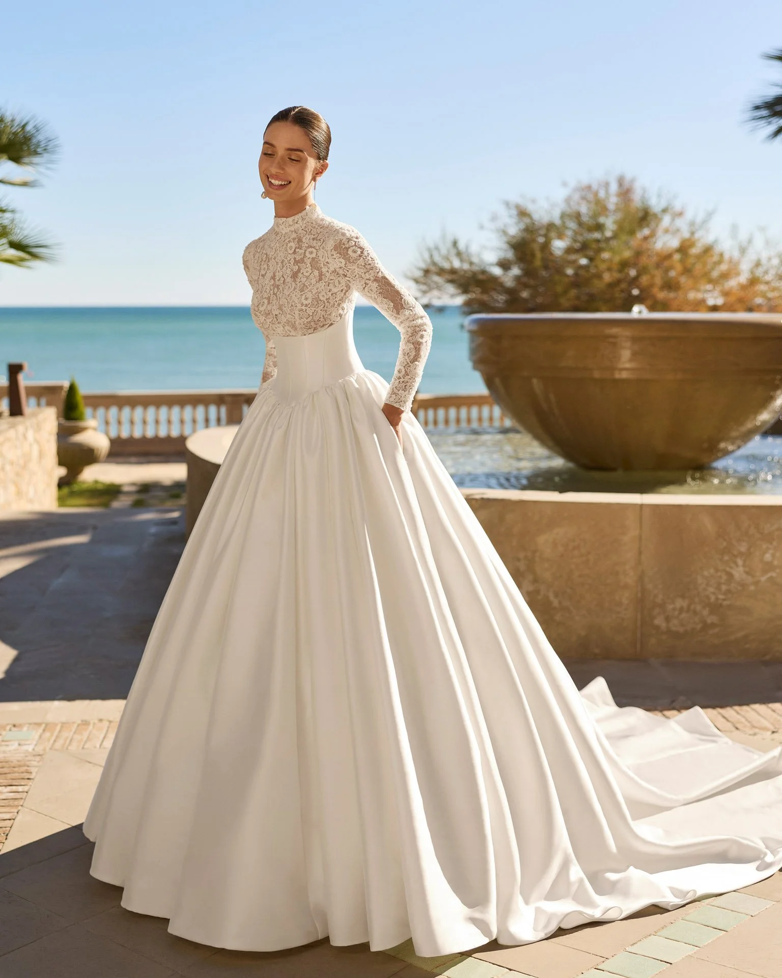 Vestido de noiva Frisa de corte princesa, com cintura basca definida, renda Chantilly e rebordé bordada, gola, mangas compridas e saia volumosa com pregas.