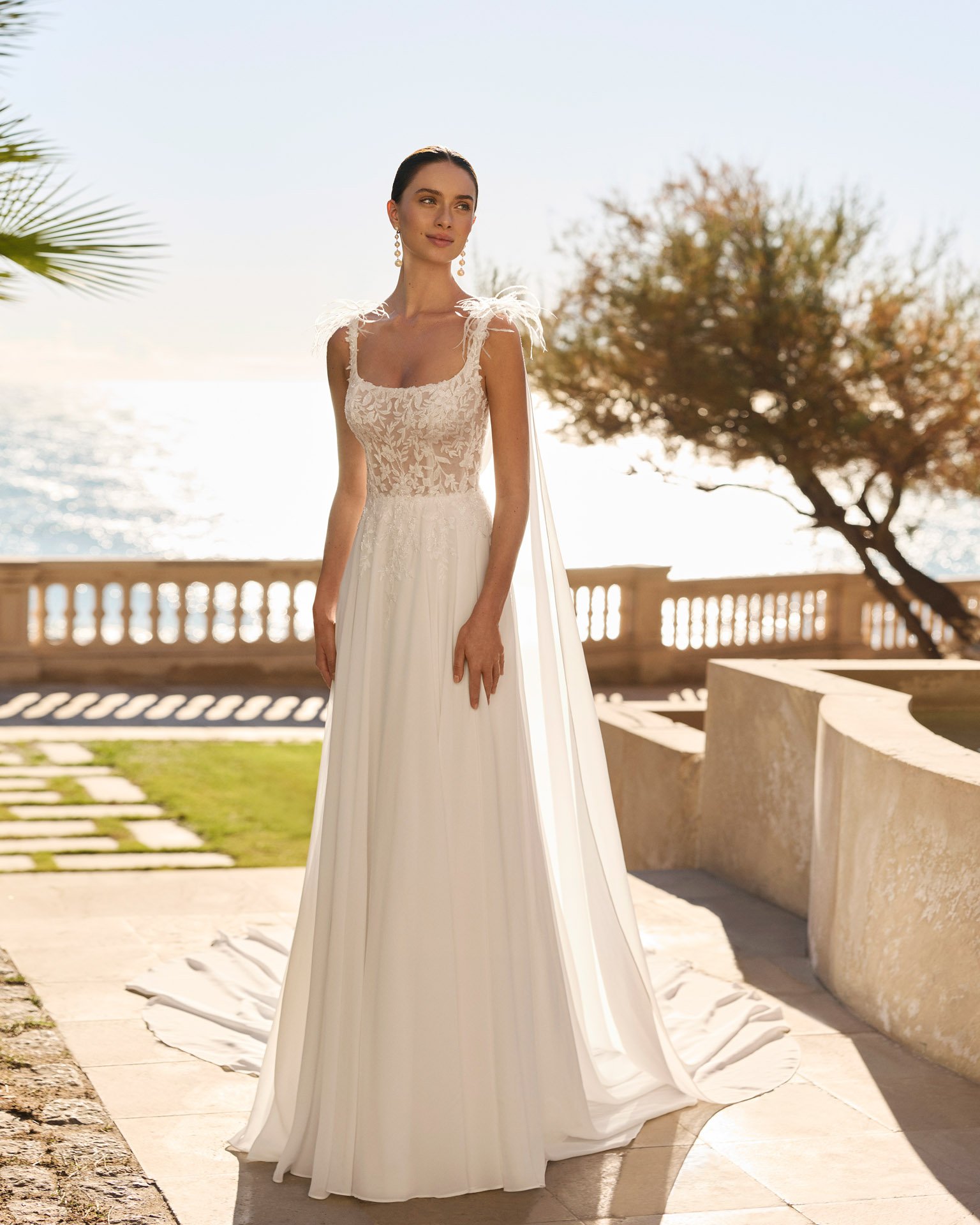 Vestido de noiva Feliz de corte esvoaçante em georgette, com decote quadrado, alças finas, corpete rendado e bordado com pedrarias e plumas nos ombros.