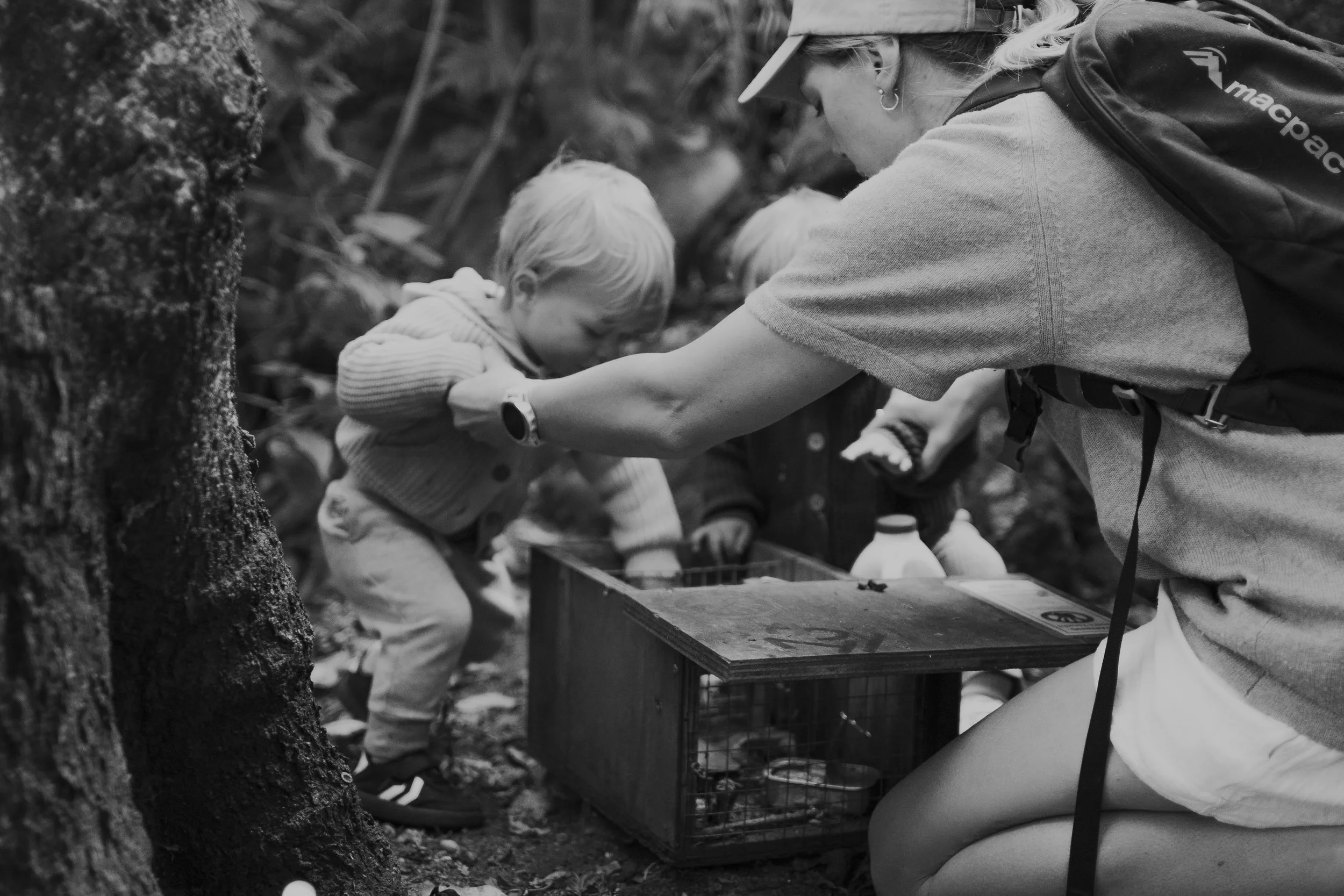 Young child playing outdoors, holding a large white plastic container over their head, wearing a gray knit sweater and gray pants, on a dirt path surrounded by greenery.