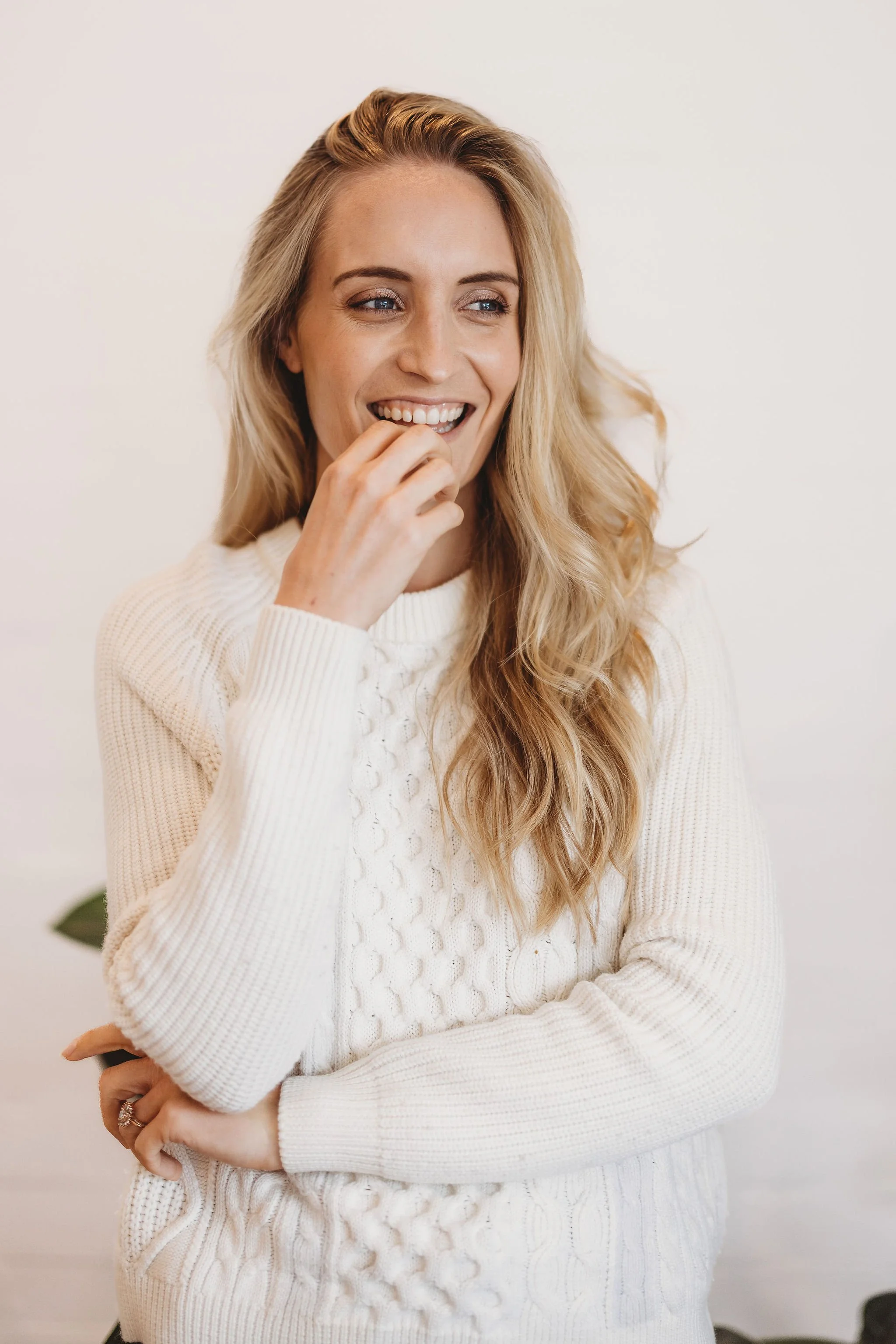A woman with long blonde hair smiling, wearing a white knitted sweater, standing against a plain white background.