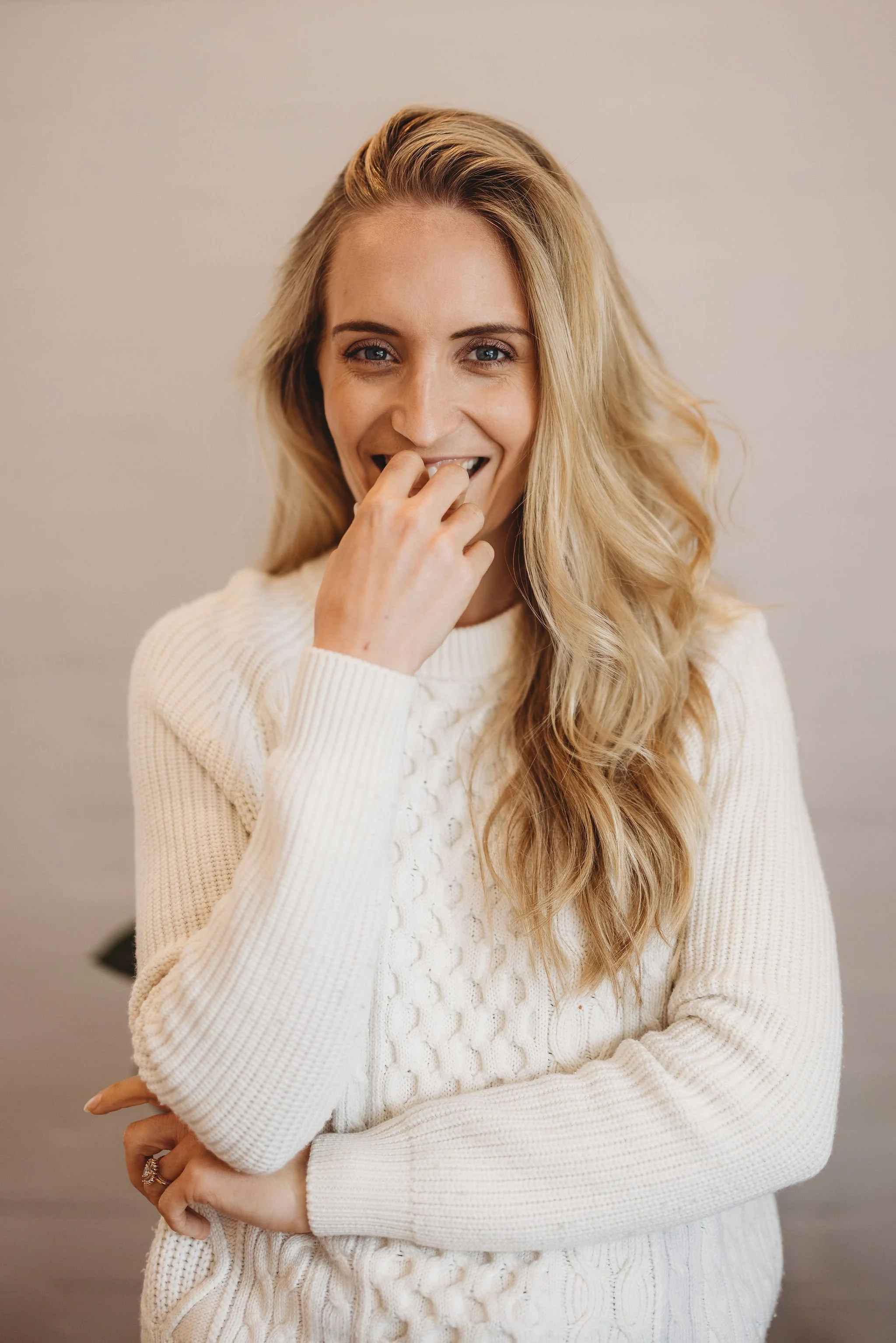 A woman with long blonde hair, wearing a white knit sweater, smiling and touching her face with her right hand.
