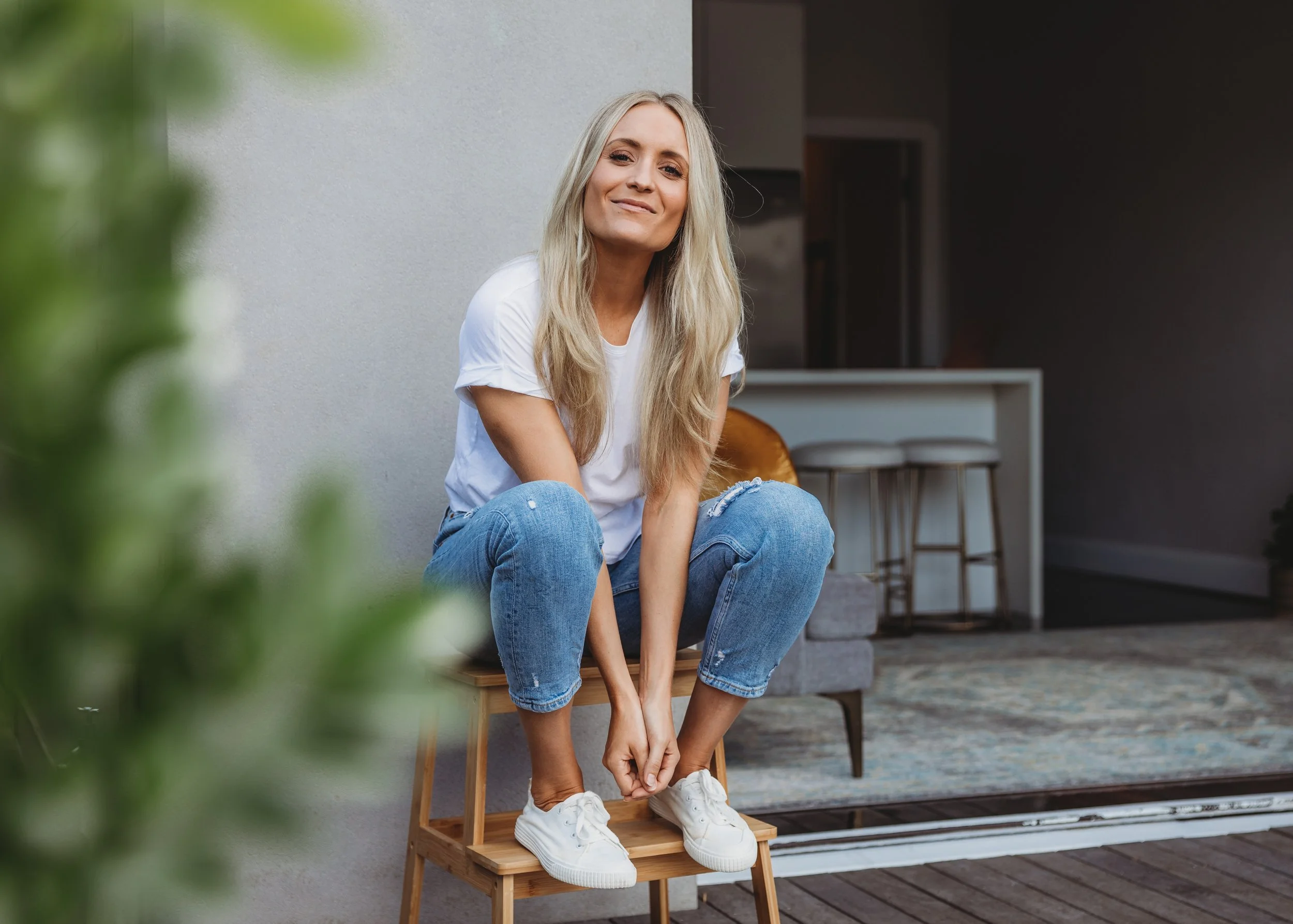 A woman with long blonde hair, wearing a white t-shirt, blue jeans, and white sneakers, sitting on a small wooden stool on a porch, smiling at the camera.