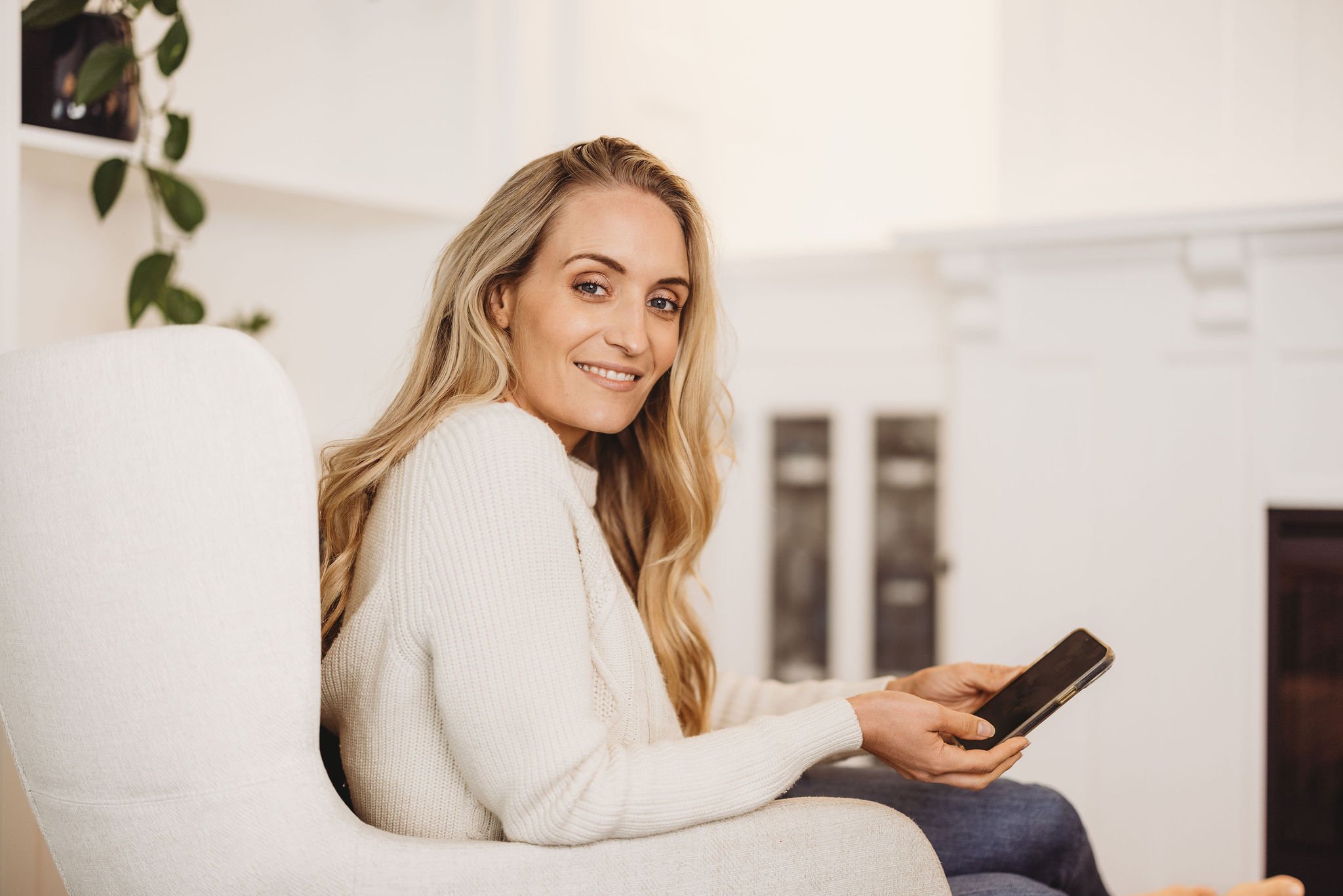 A woman with long blonde hair sitting on a white chair, smiling at the camera, holding a smartphone in her hands, in a bright room with white walls and a fireplace.