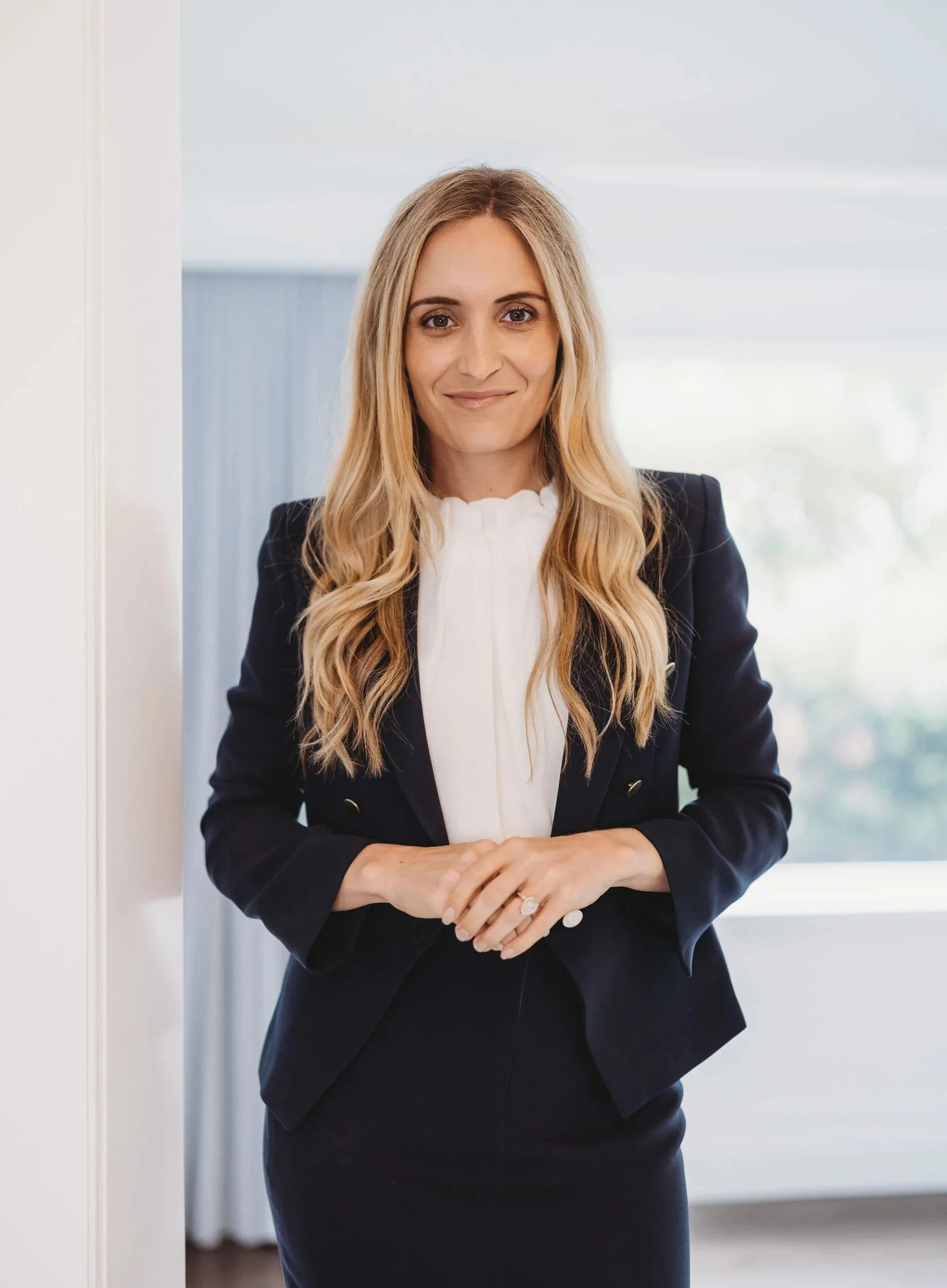 A professional woman with long blonde hair wearing a navy blazer and white blouse standing indoors near a window.