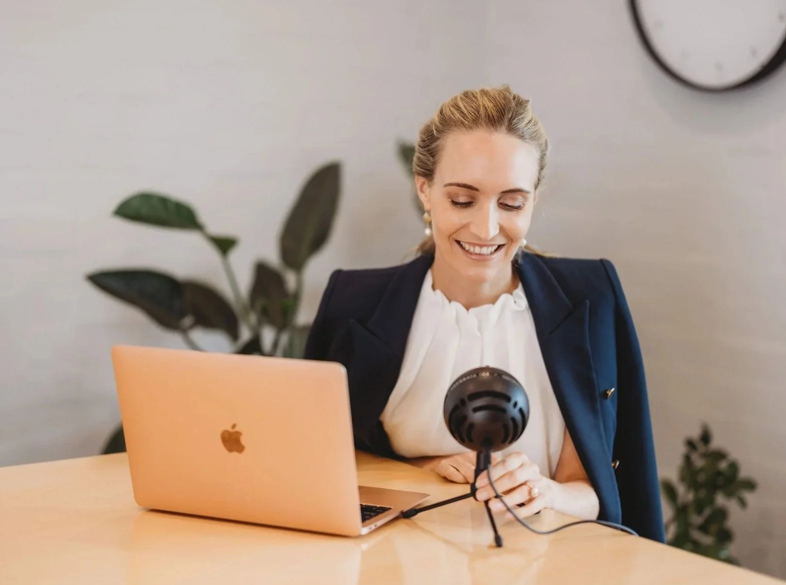 A woman sitting at a desk with a laptop and a microphone, smiling and looking down.
