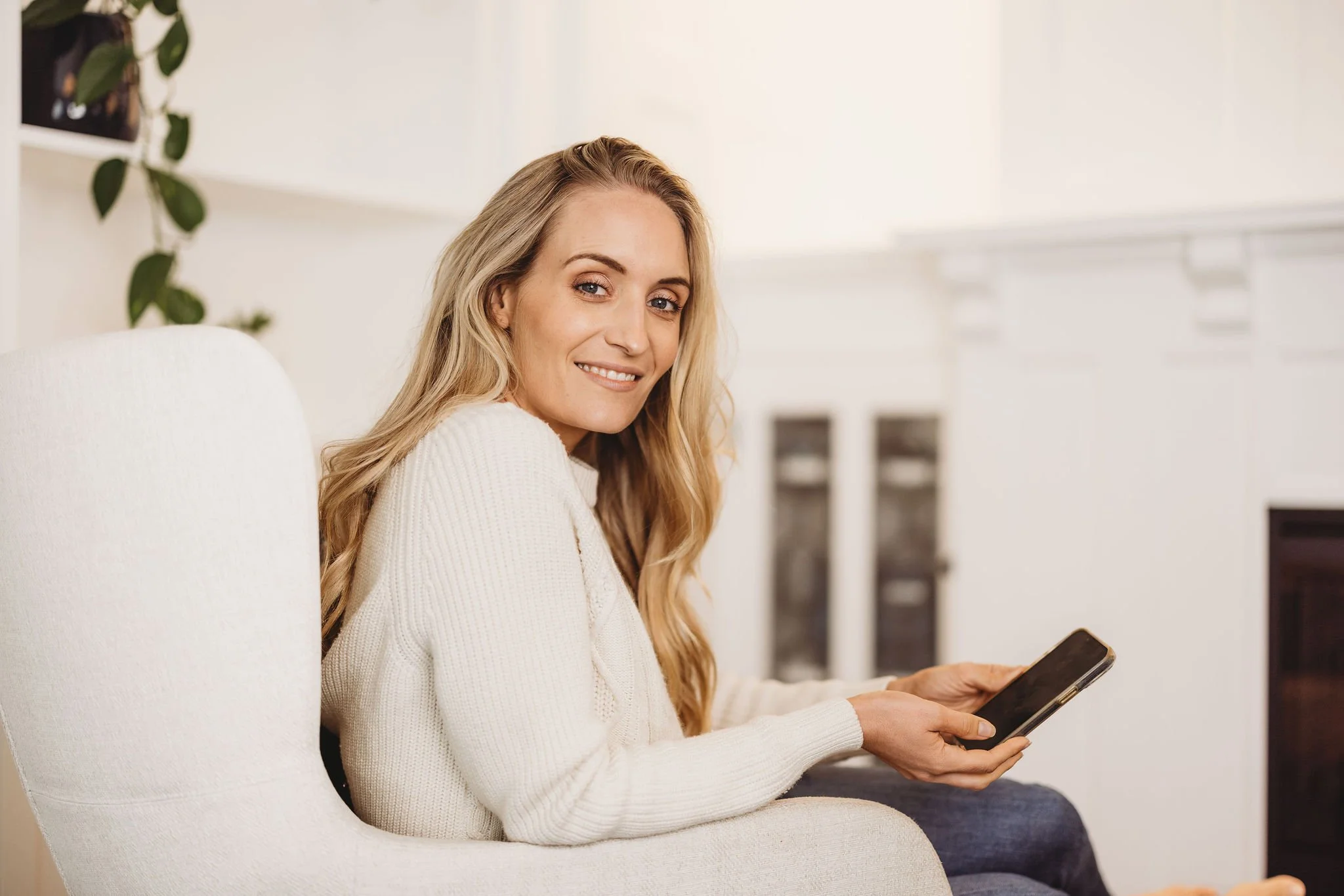A woman with long blonde hair sitting on a white armchair, smiling, holding a smartphone in her hand in a bright room.