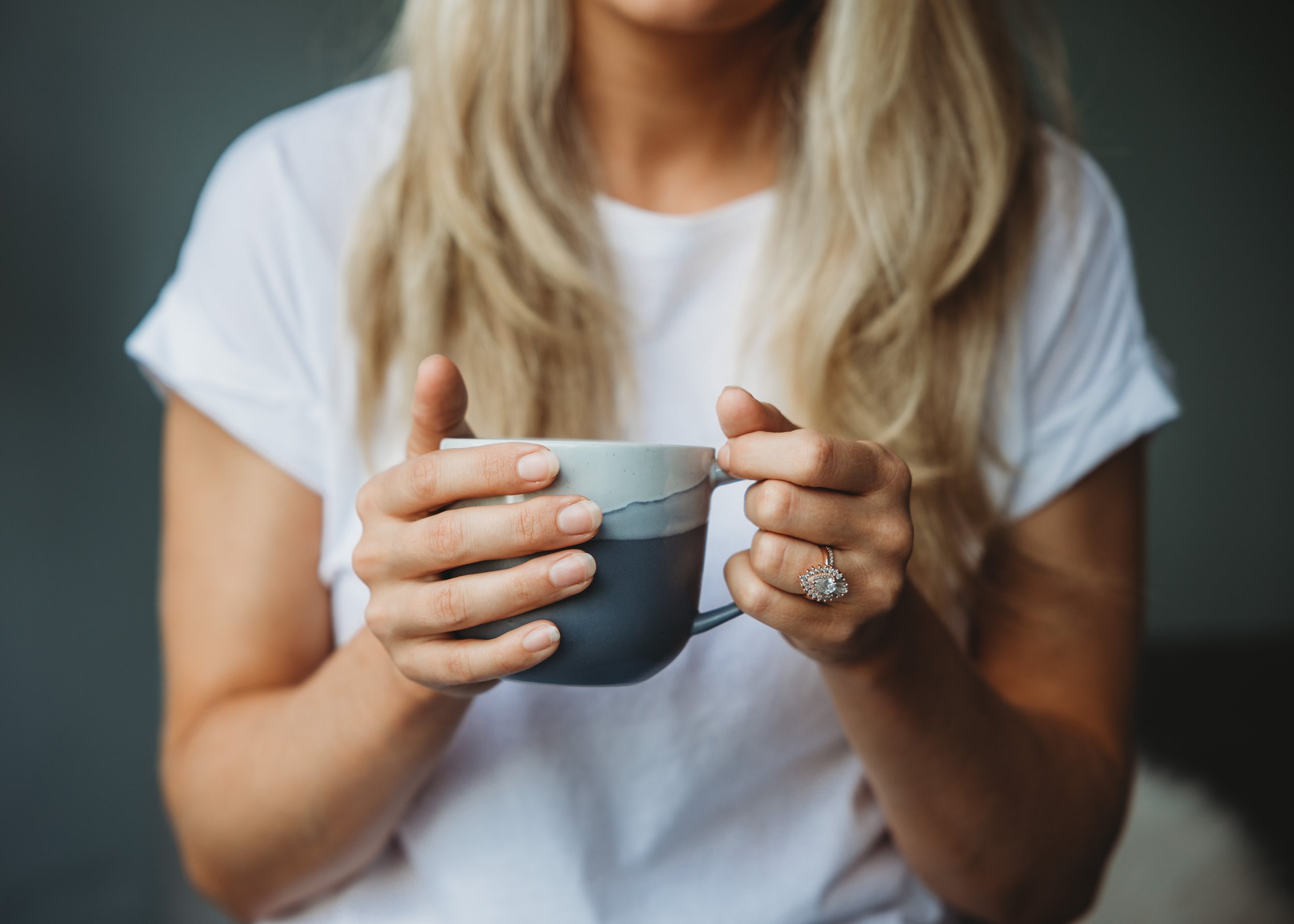 A woman with blonde hair wearing a white t-shirt holding a ceramic mug with both hands.