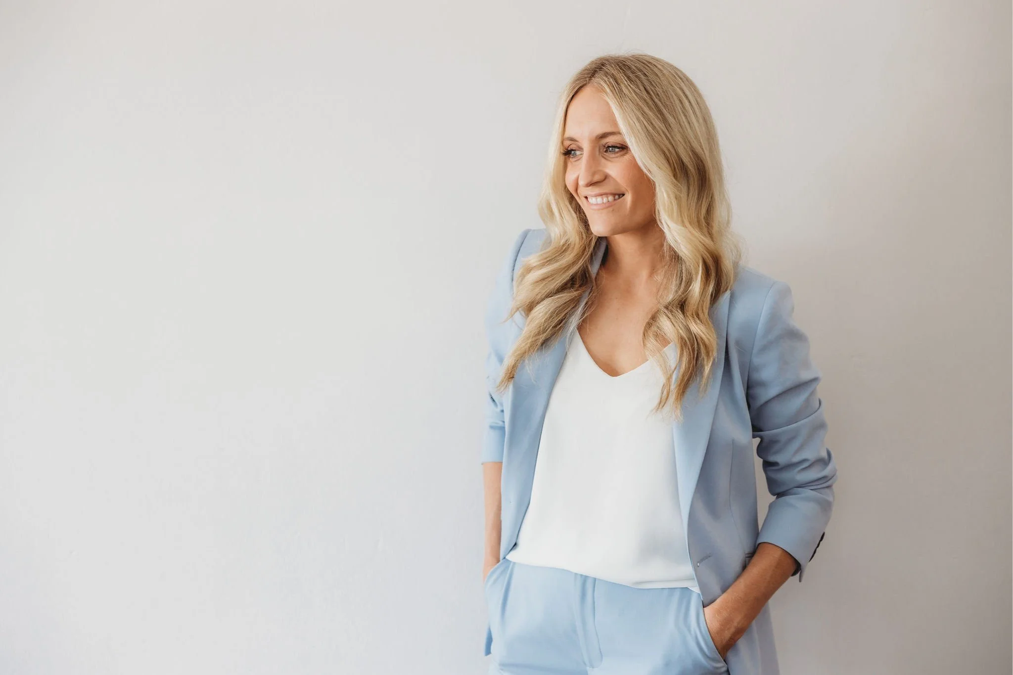 A woman with long blonde hair wearing a light blue suit and white top, smiling and standing against a plain white wall.