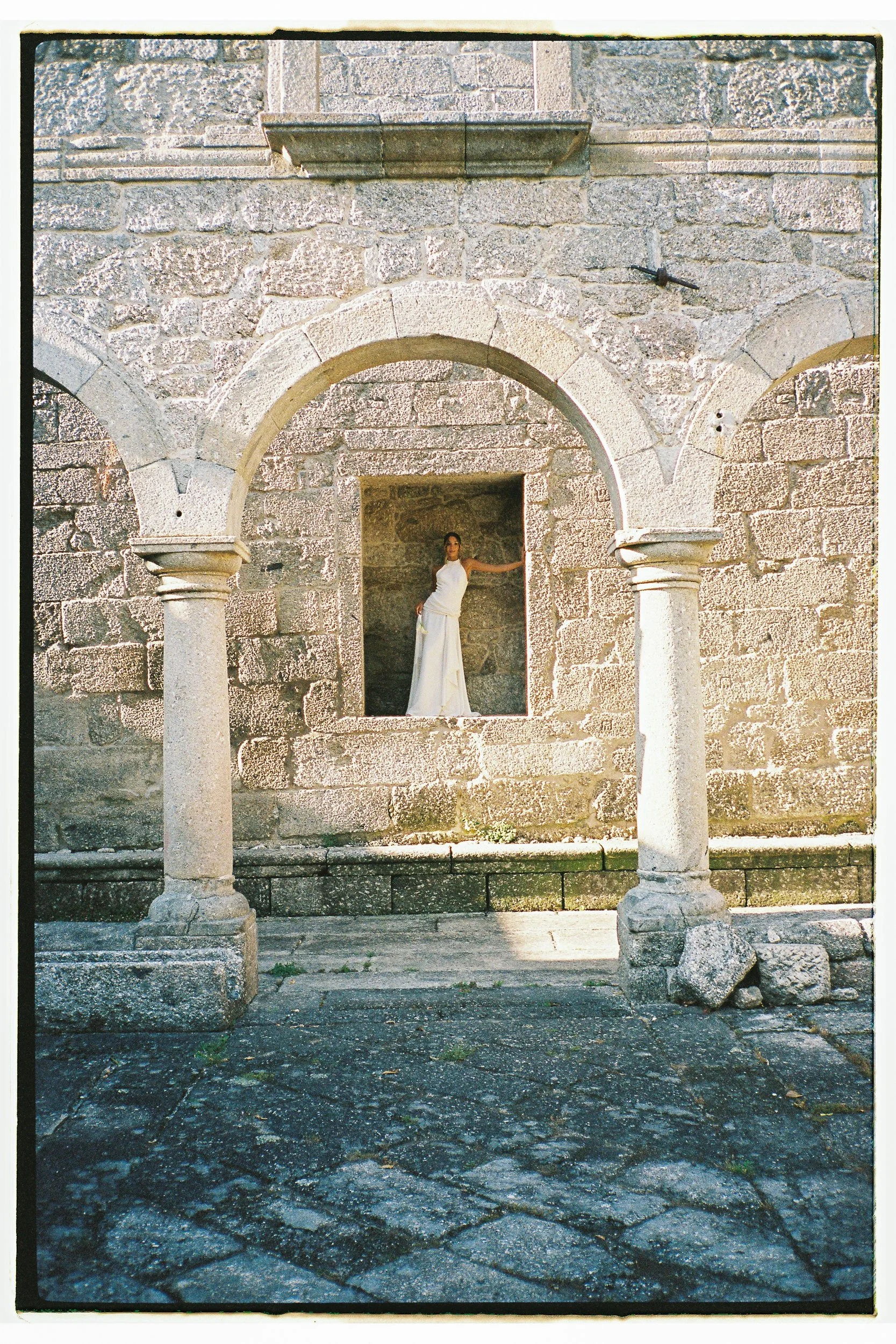 A woman in a white dress standing under Roman-style stone arches of an old stone building, with stone paving ground.