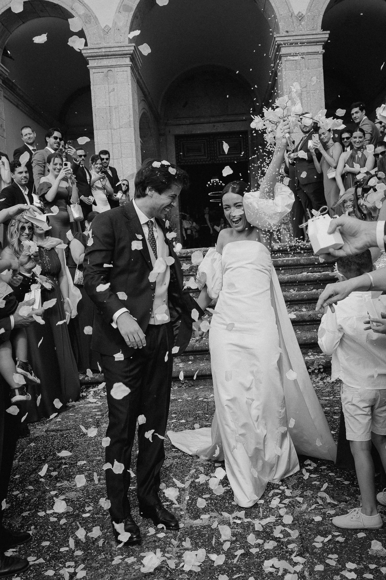 Black and white photo of a wedding celebration outside a building with large columns. The bride and groom are smiling and laughing as they walk on scattered confetti, surrounded by friends and family taking photos and videos.