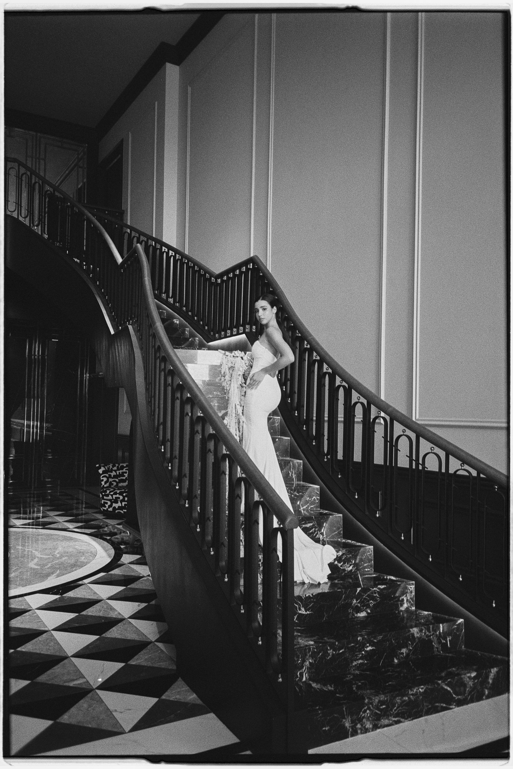 Black and white photo of a woman in a strapless dress standing on a staircase with a black metal railing, in an elegant interior with patterned flooring and tall wall panels.