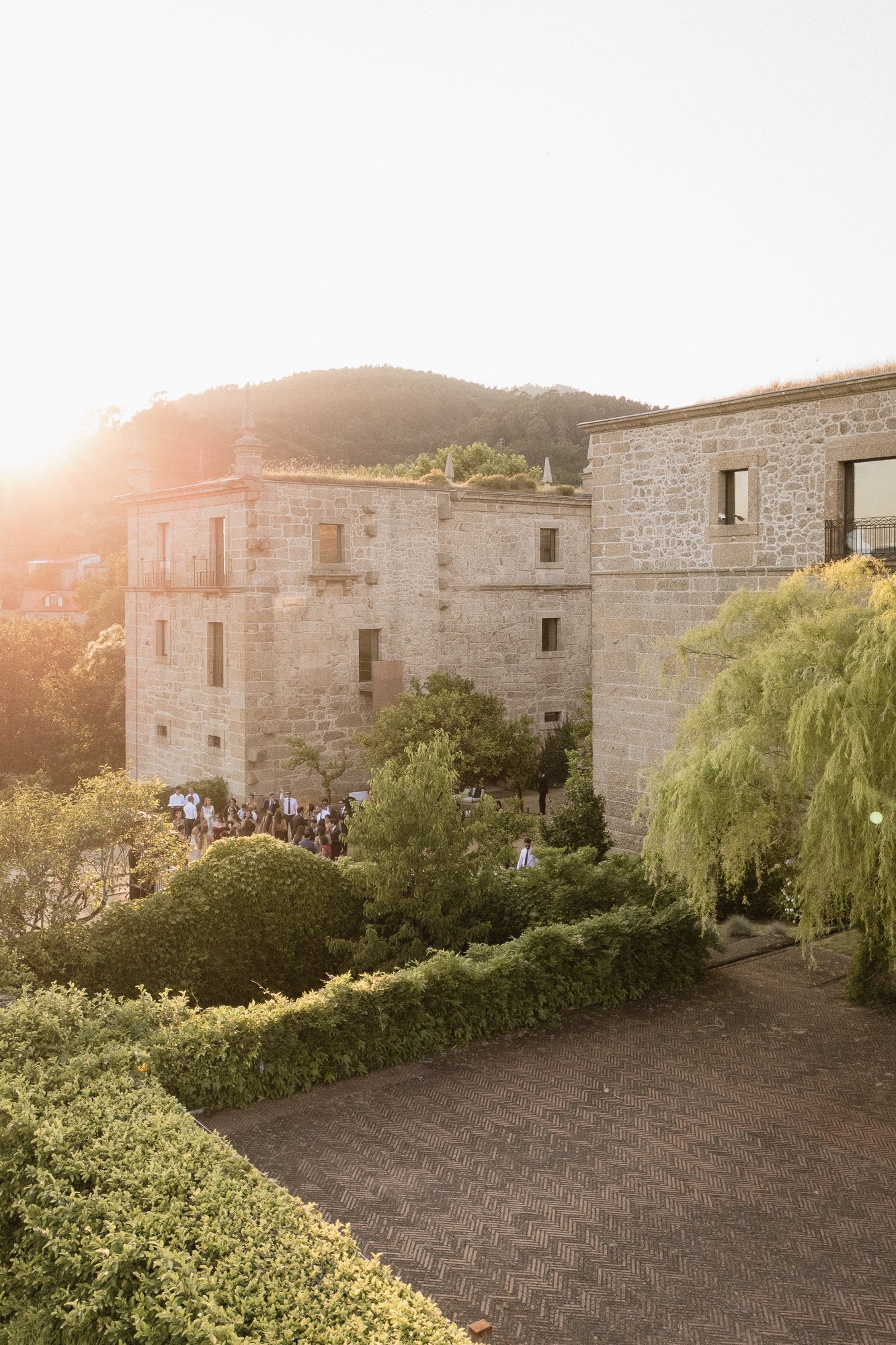 A stone castle-like building with multiple windows and small balconies, set in a lush garden with trees and bushes, under bright sunlight during sunset or sunrise.