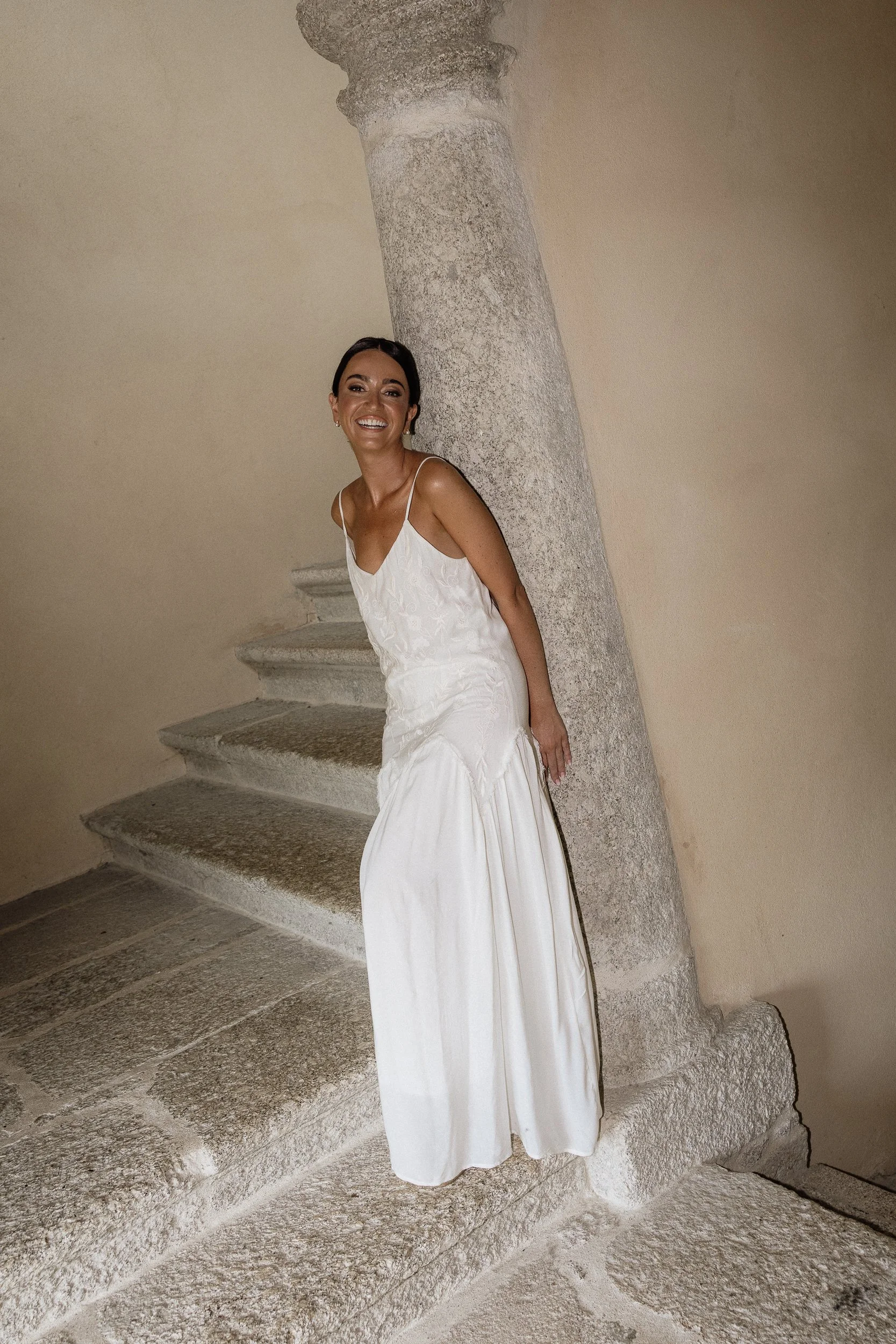 Woman in a white dress standing on stone stairs next to a large stone column, smiling at the camera.