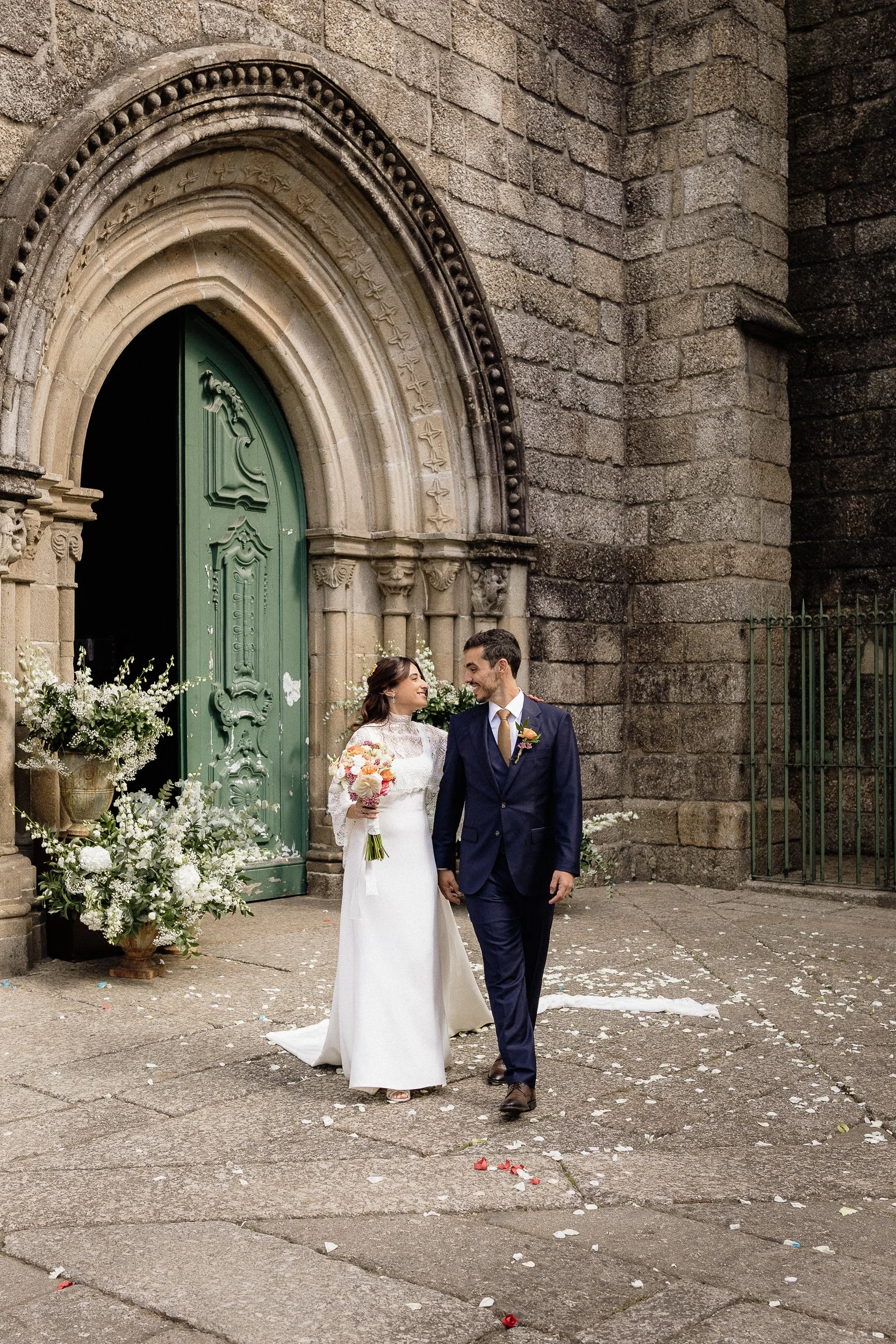 A bride and groom standing outside a stone church, holding hands and smiling at each other. The bride is holding a bouquet of flowers, and both are dressed in wedding attire. There are flower arrangements and flower petals on the ground, with an old stone wall and green church door in the background.