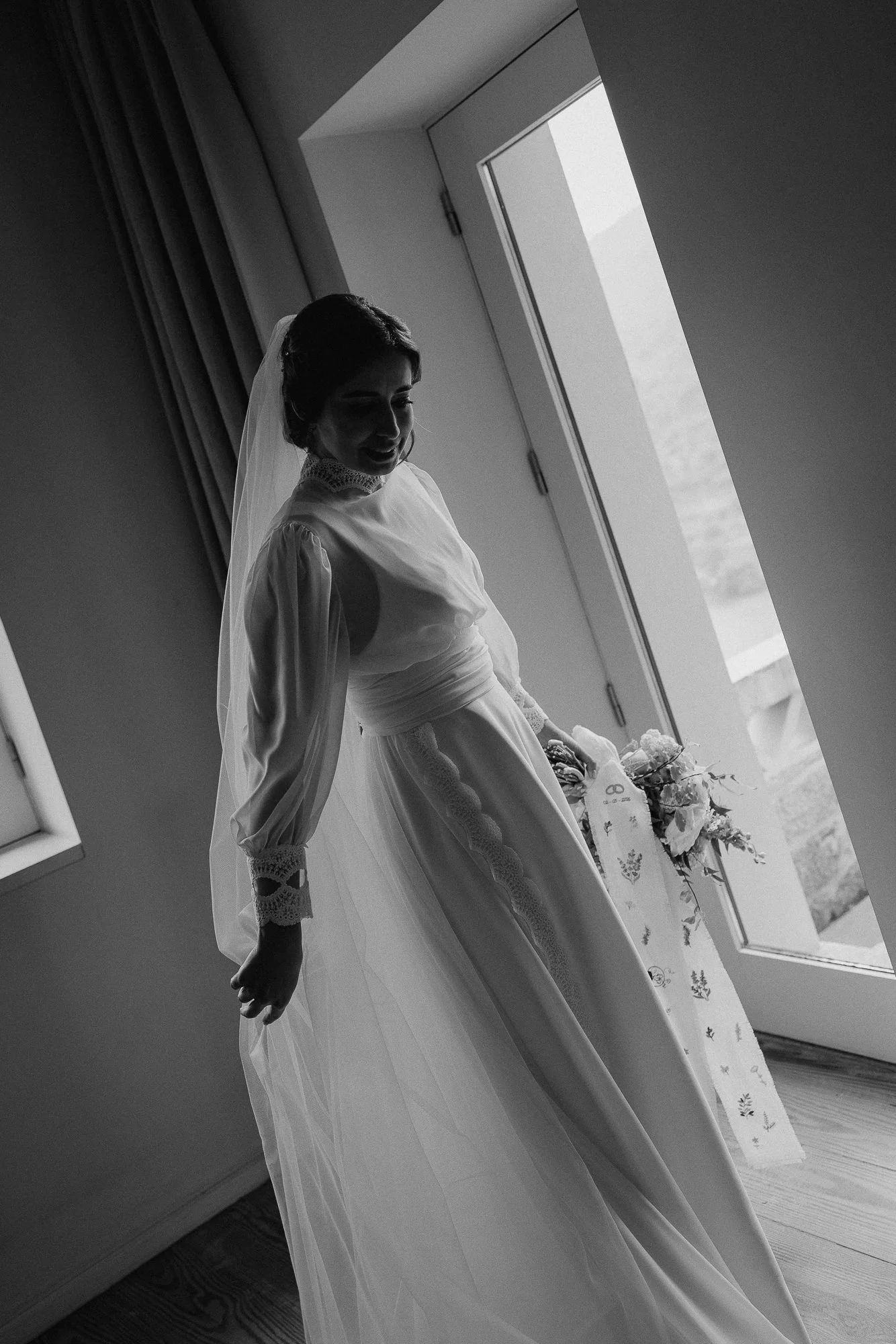 Black and white photo of a bride standing by a window, smiling and looking down, with her hair styled short and wearing a wedding dress with lace details and a veil, in a softly lit interior.