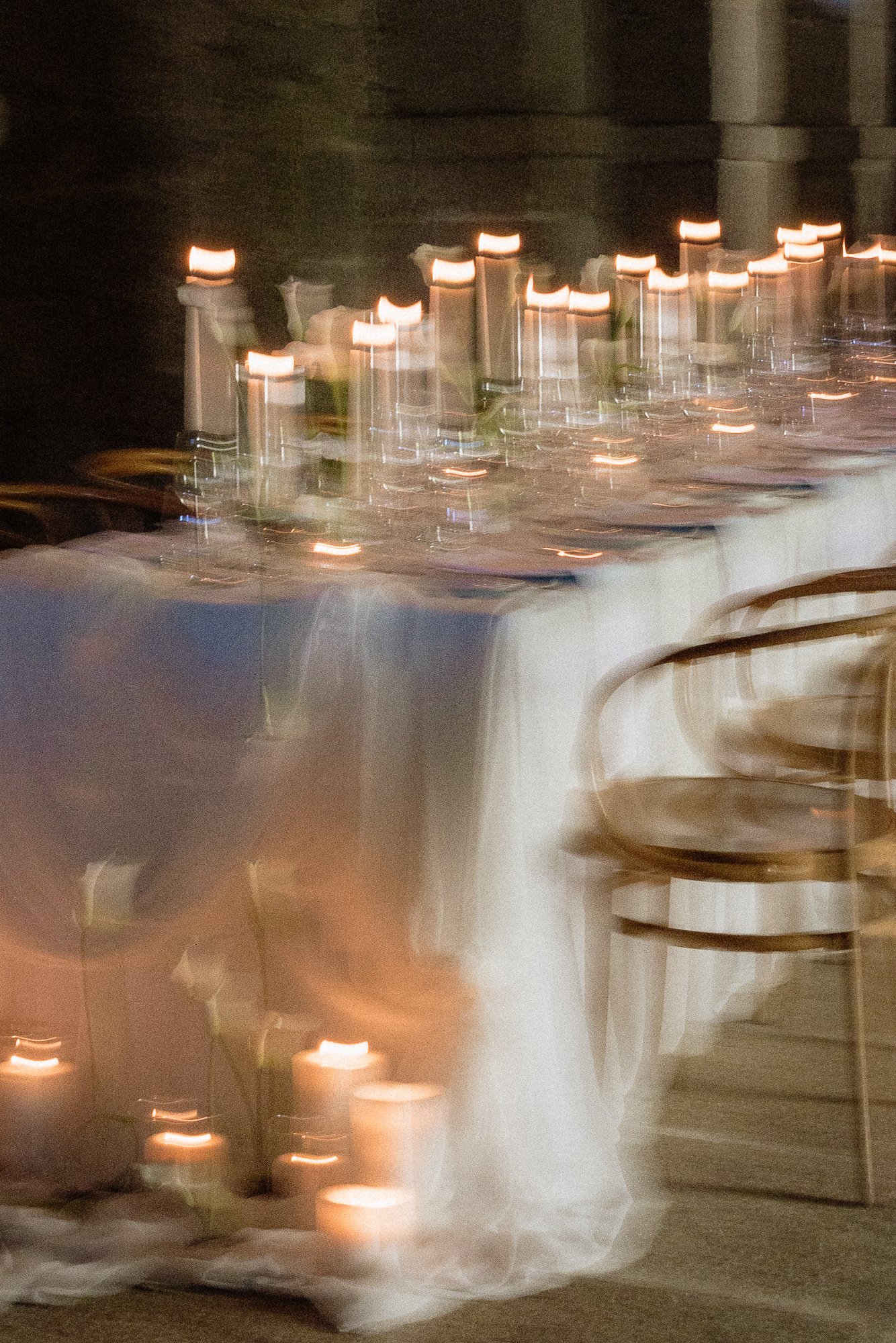 Blurred image of a row of lit candles on a dark background, with seating chairs in the foreground.