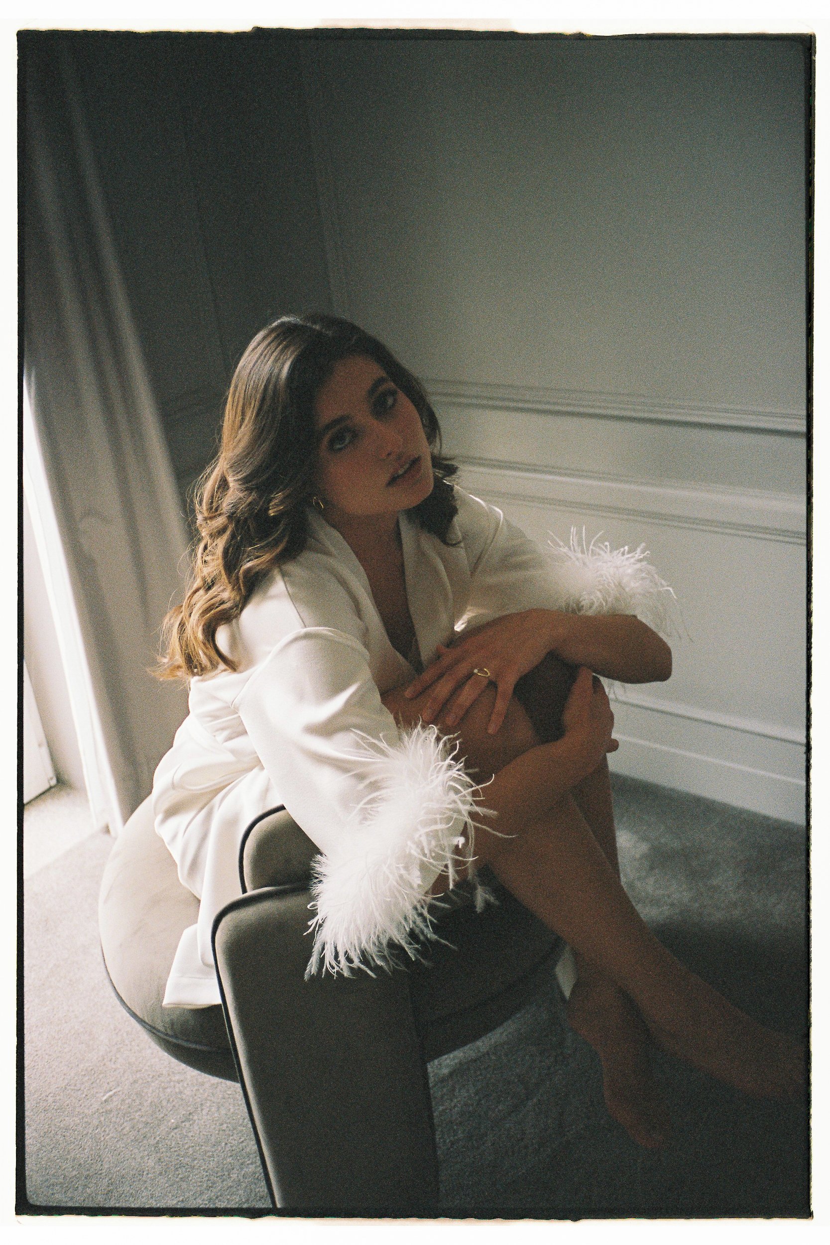 A woman with long wavy hair and makeup sitting on a chair in a room with a white wall and blinds, wearing a white satin robe with feathered cuffs, holding her leg, and looking at the camera.