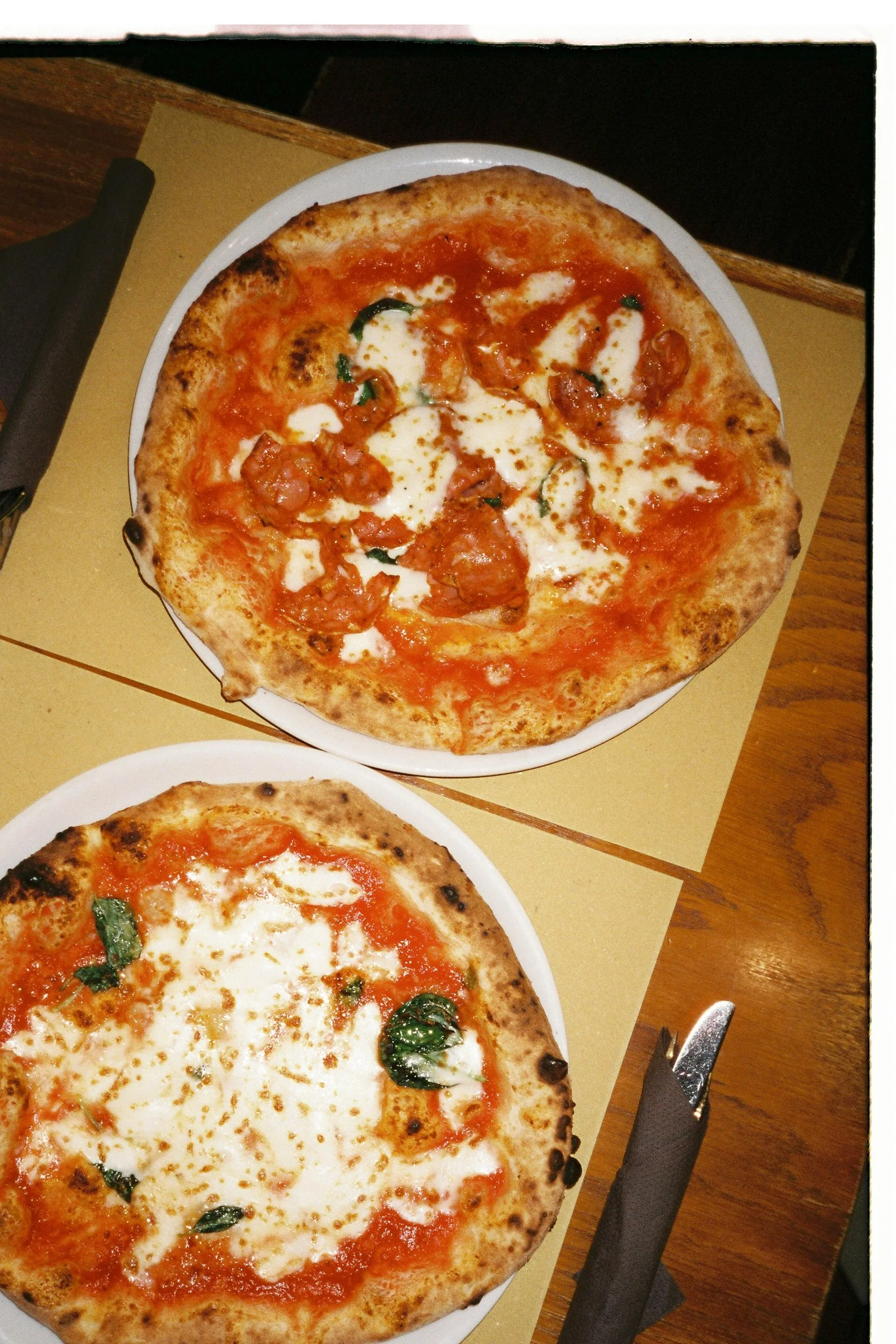 Close-up of two Margherita pizzas on white plates, with melted cheese, tomato sauce, and basil leaves, on a table with utensils and a napkin.