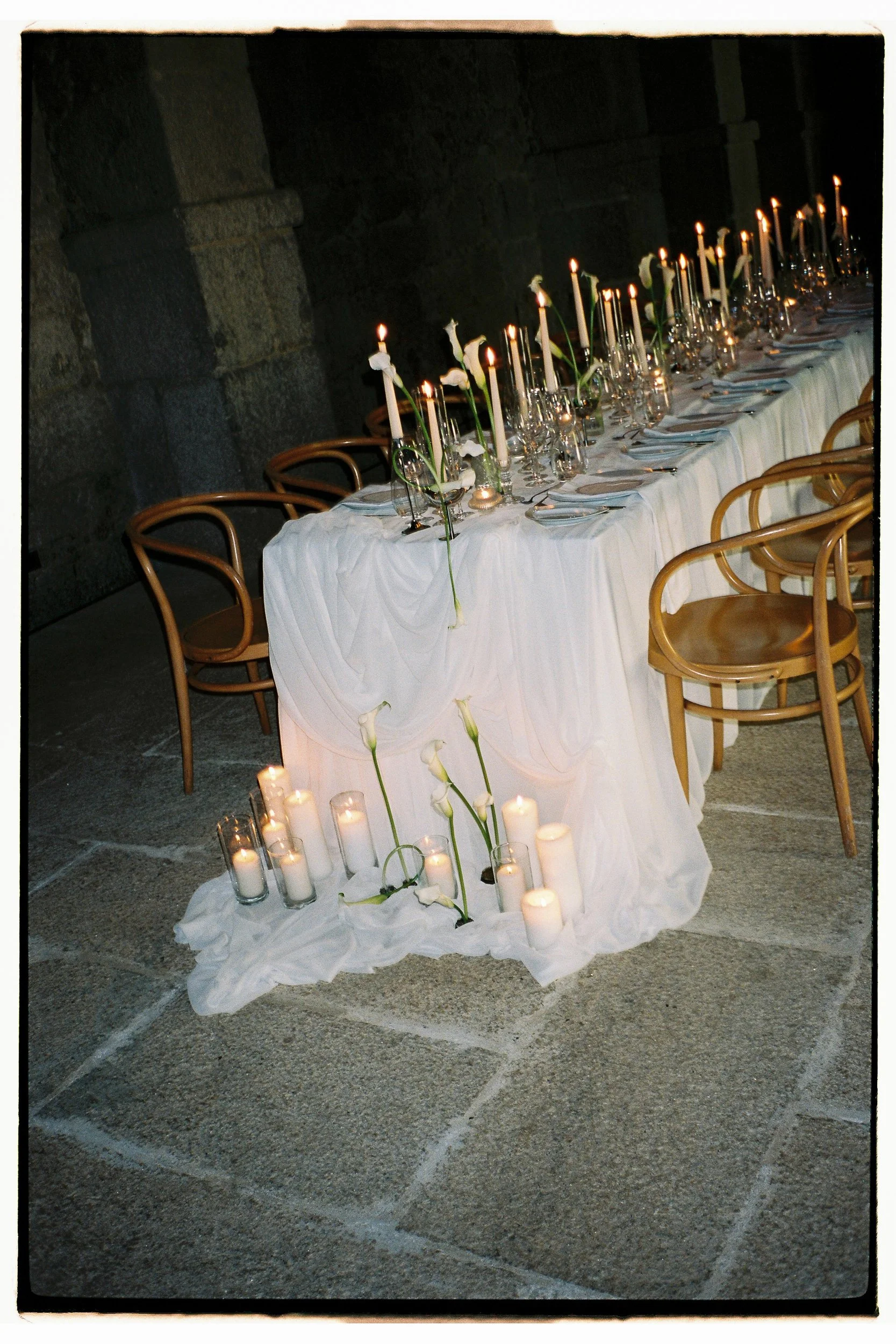 A long table with white tablecloth, decorated with white candles and white flowers, set for a formal event or dinner in a dimly lit stone-walled room.
