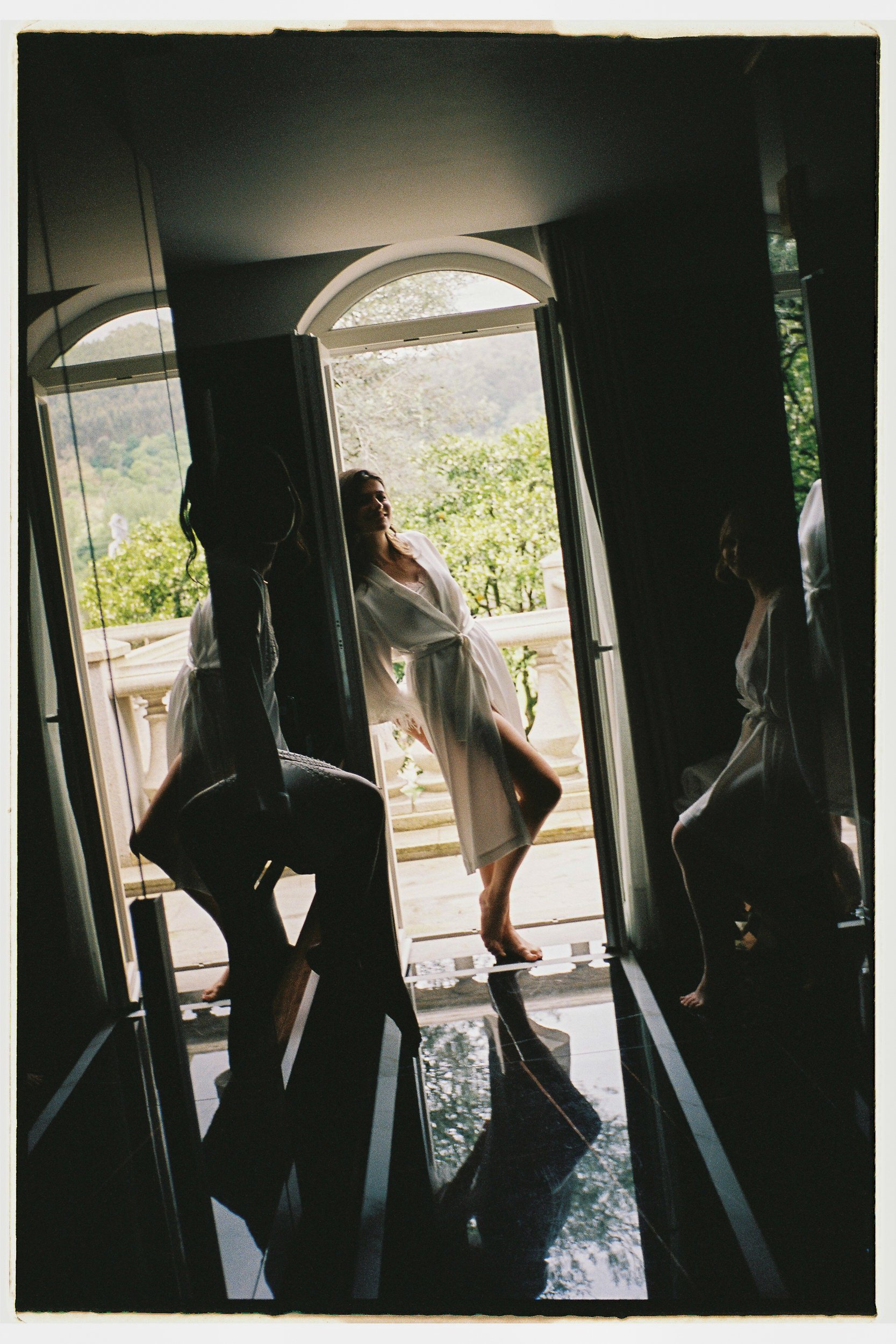 Women in satin robes standing barefoot on a black tiled floor, reflected in a large mirror, with open doors showing a balcony and greenery outside.