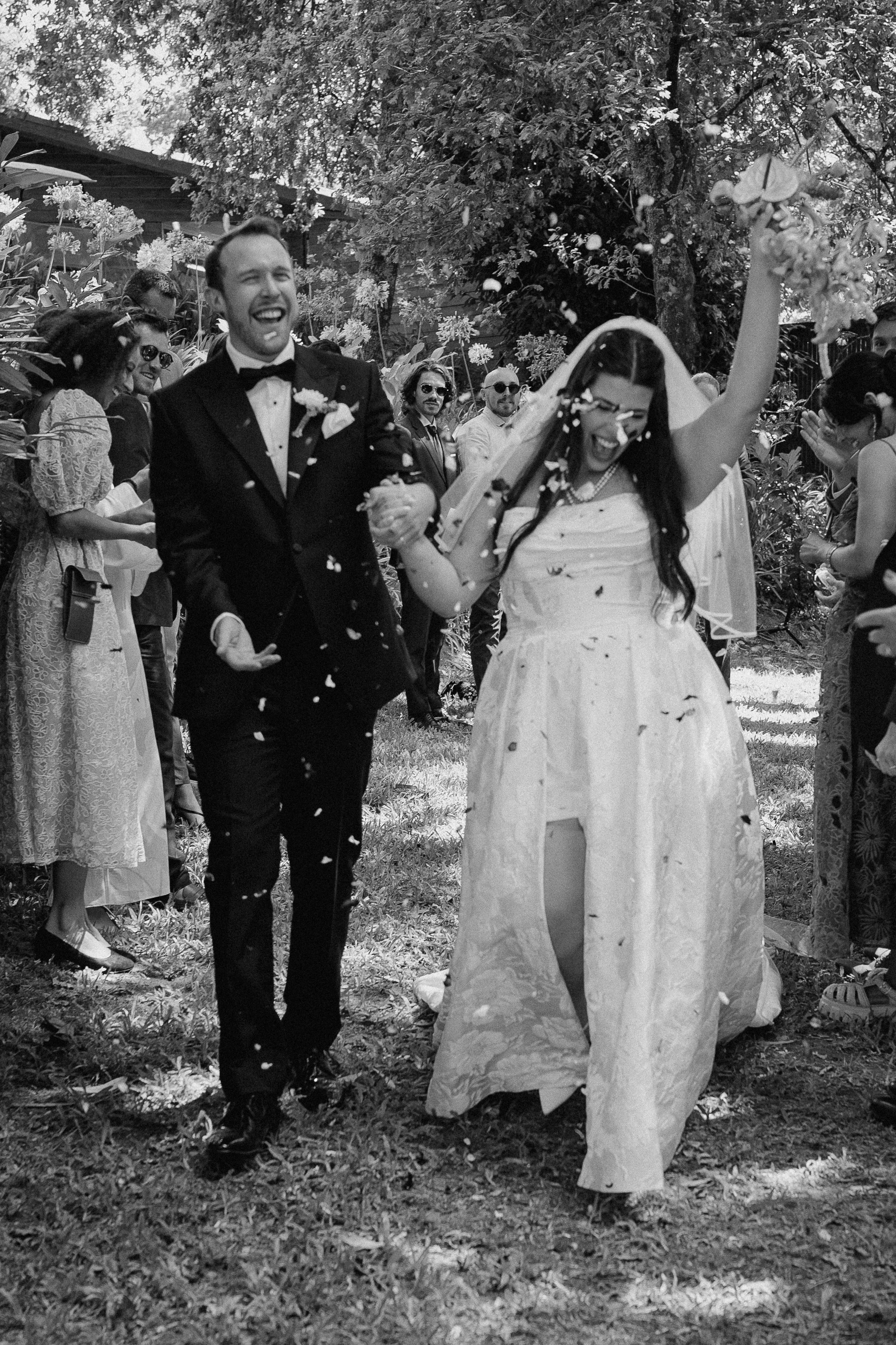 A black and white photo of a wedding celebration outdoors, featuring a groom in a tuxedo and a bride in a wedding gown, smiling and holding hands with children, surrounded by guests and trees.