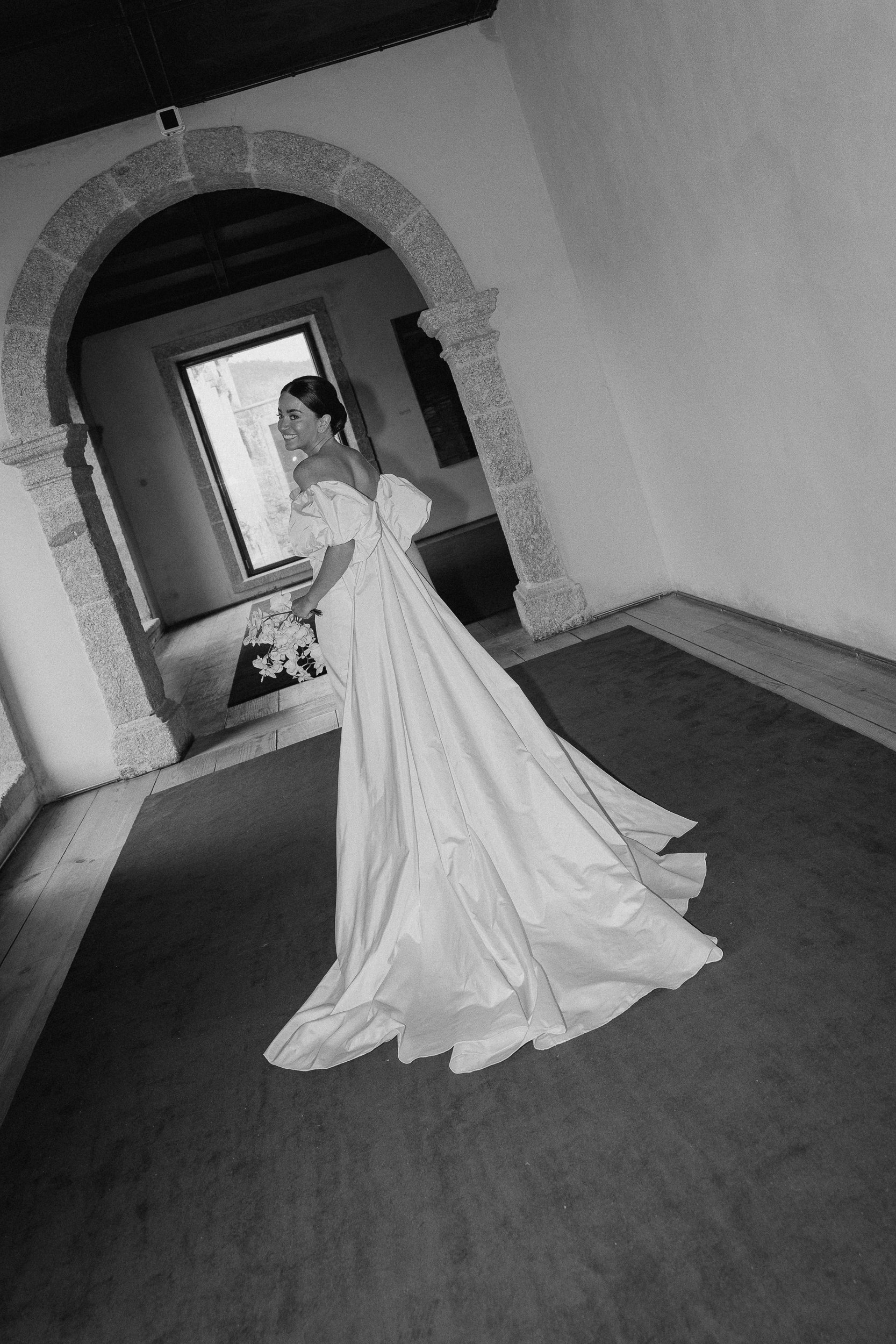 A woman in a wedding dress smiling and holding a bouquet, standing in a room with stone archways and wooden flooring, black and white photograph.