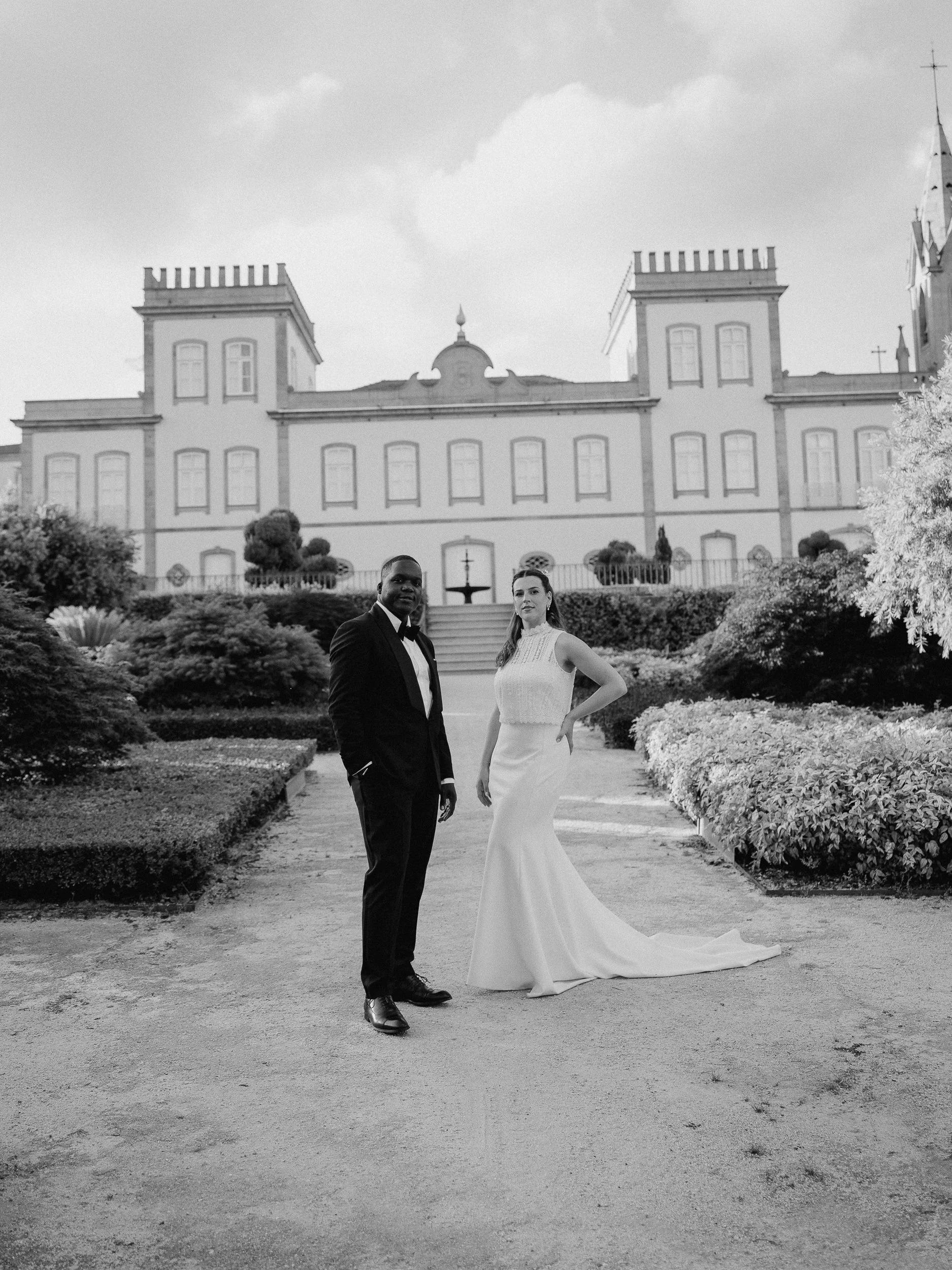Black and white photo of a bride and groom standing on a path in front of a large estate or castle, surrounded by landscaped gardens and bushes.