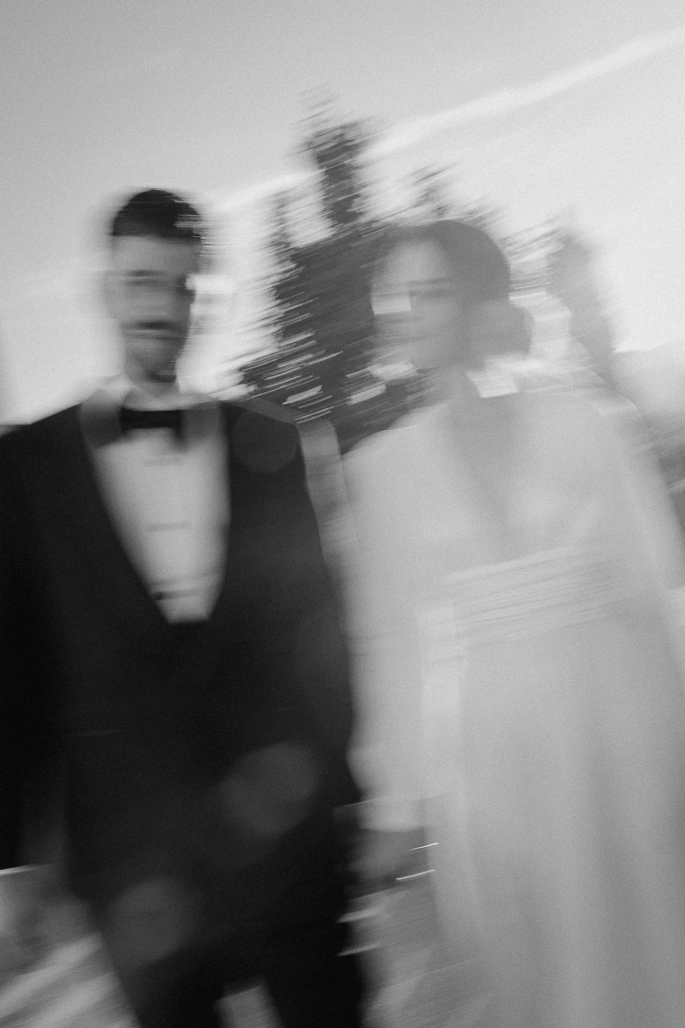Black and white photo of three people in formal attire, blurred due to motion, with one person prominently in the foreground and two in the background.