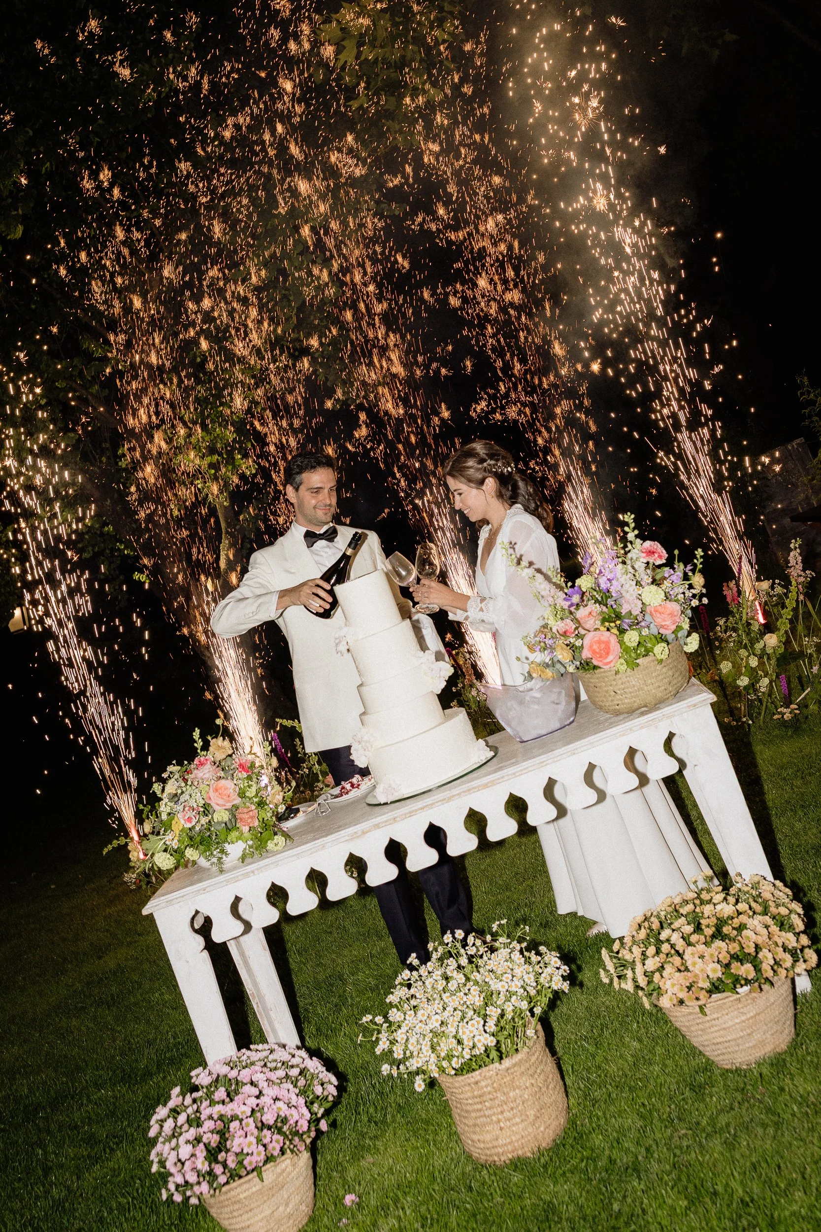 A couple celebrating wedding with a white cake, fireworks, and flowers in the evening outdoors.